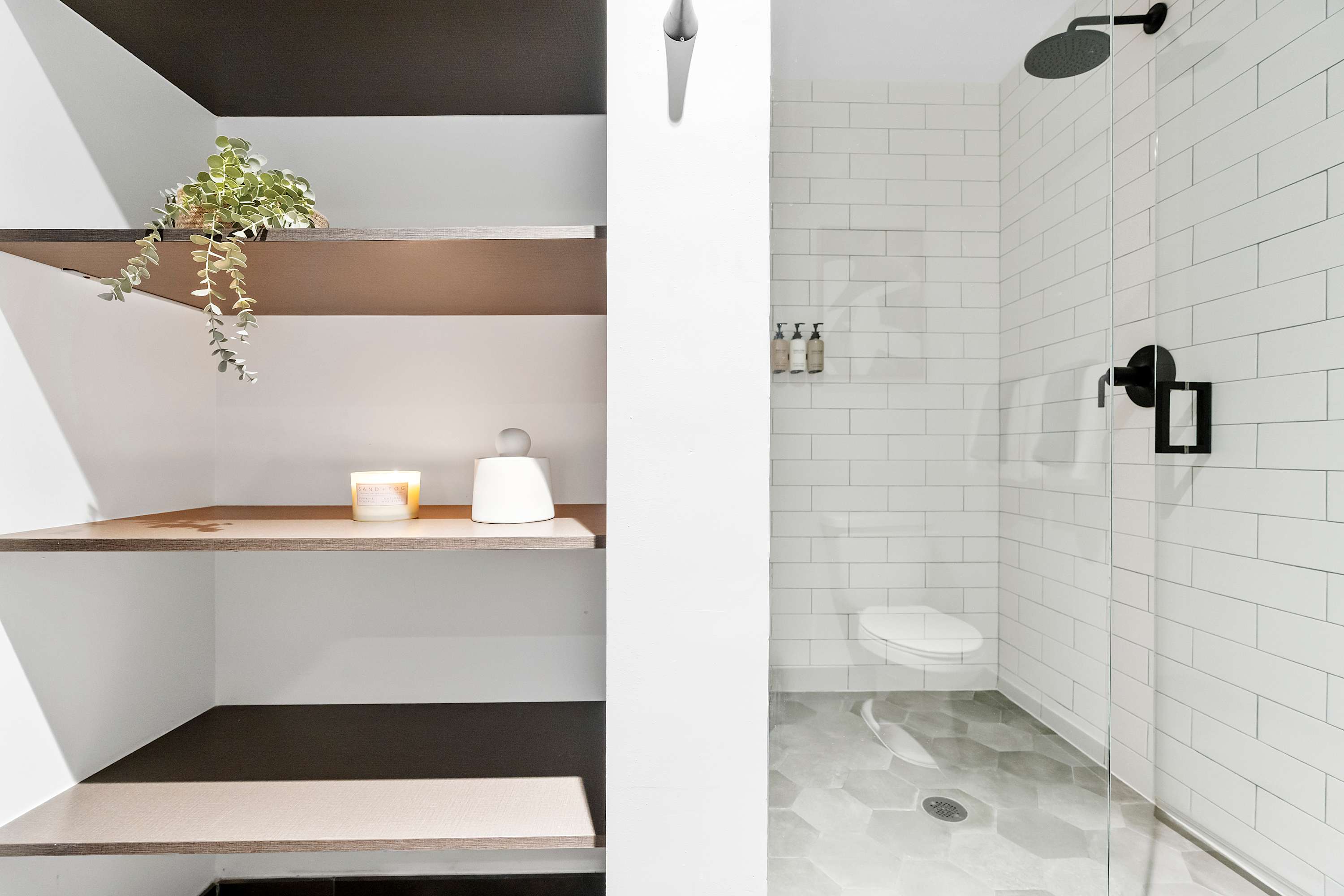 A modern walk-in shower with white subway tile walls, a glass enclosure, and a dark hexagonal tile floor, adjacent to open shelving with decorative items and a plant.