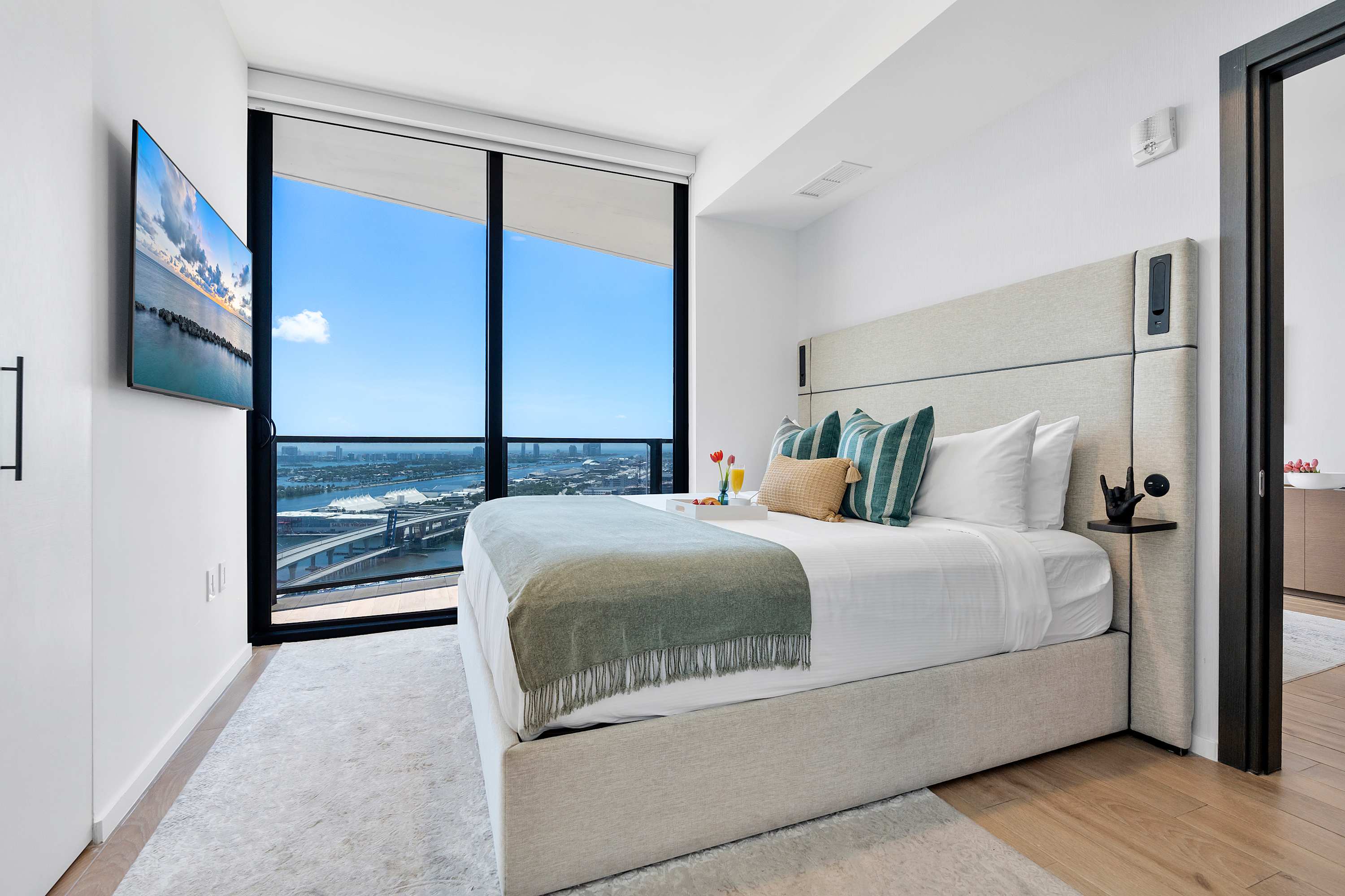 A modern bedroom with a light-colored upholstered king bed, striped accent pillows, and a large window revealing a stunning city and water vista, complemented by a light rug and wood flooring.