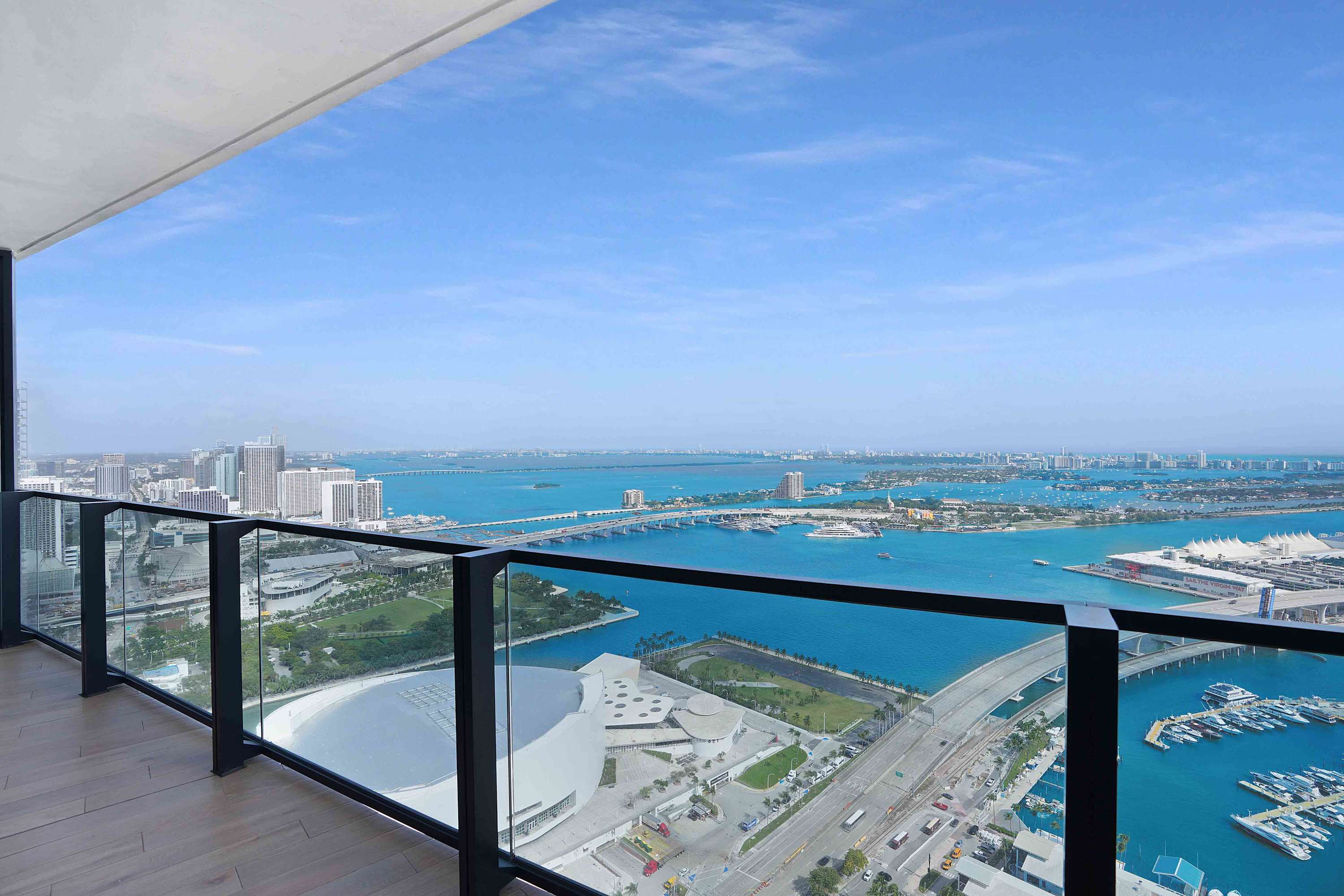 Spacious private balcony with a glass railing offers a breathtaking panoramic view of the ocean, multiple bridges, and a distant city skyline under a bright blue sky.