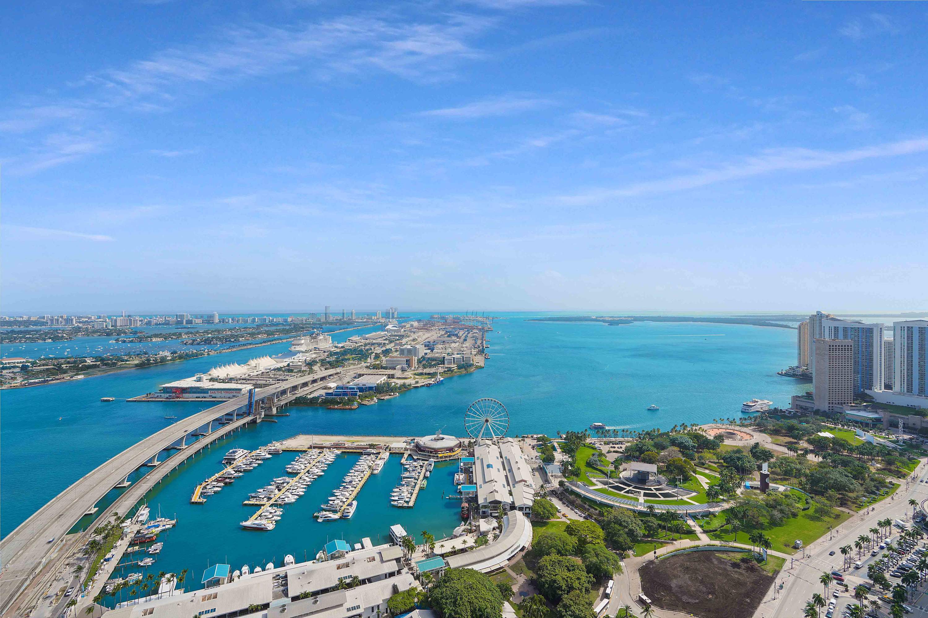 A breathtaking aerial view showcasing a vibrant city skyline, a bustling marina filled with boats, and the expansive blue ocean under a clear sky with scattered clouds.