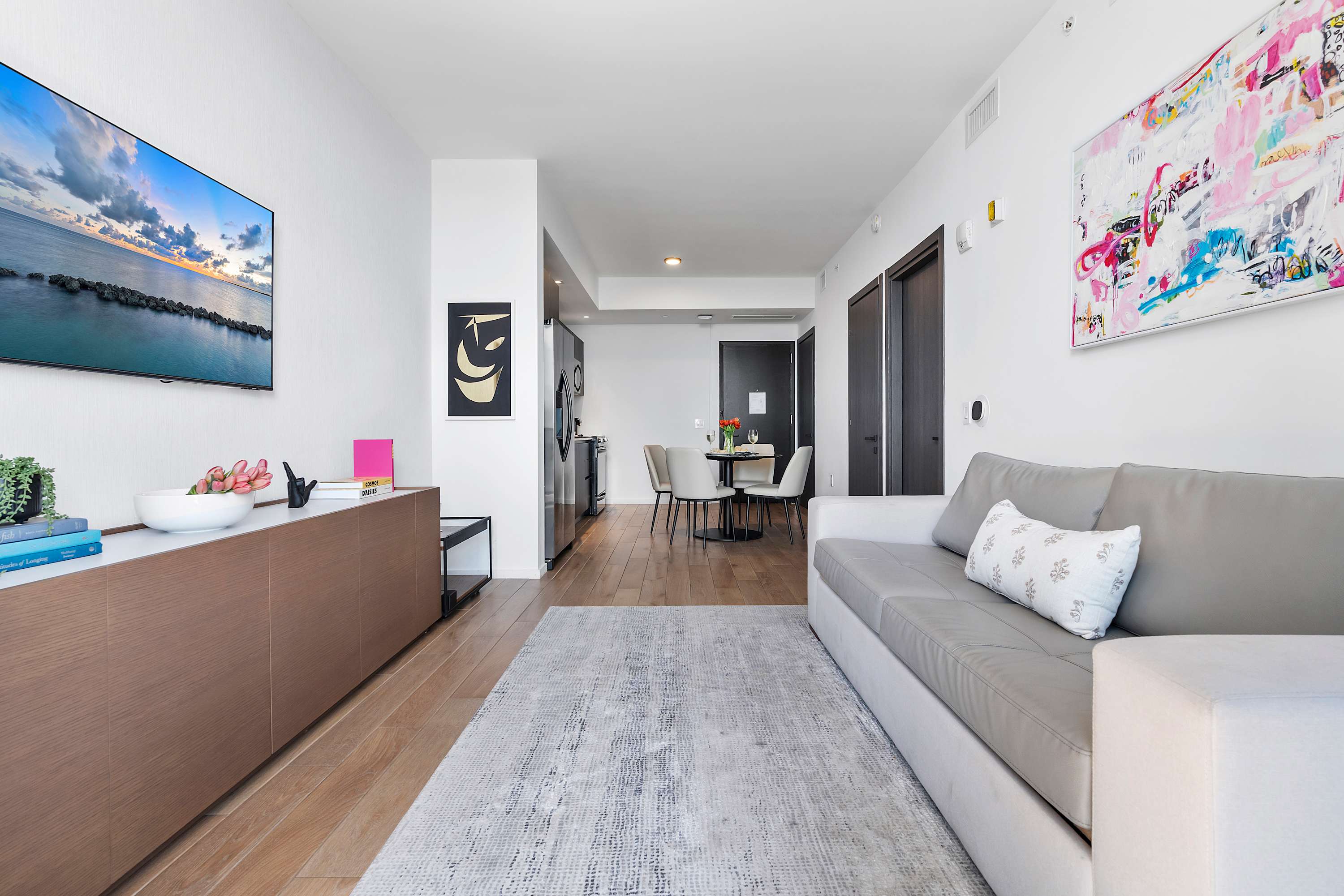 Contemporary living room features a light gray sofa, a patterned area rug, and a wall-mounted TV above a wooden console, with an open-concept view towards the dining area and kitchen.