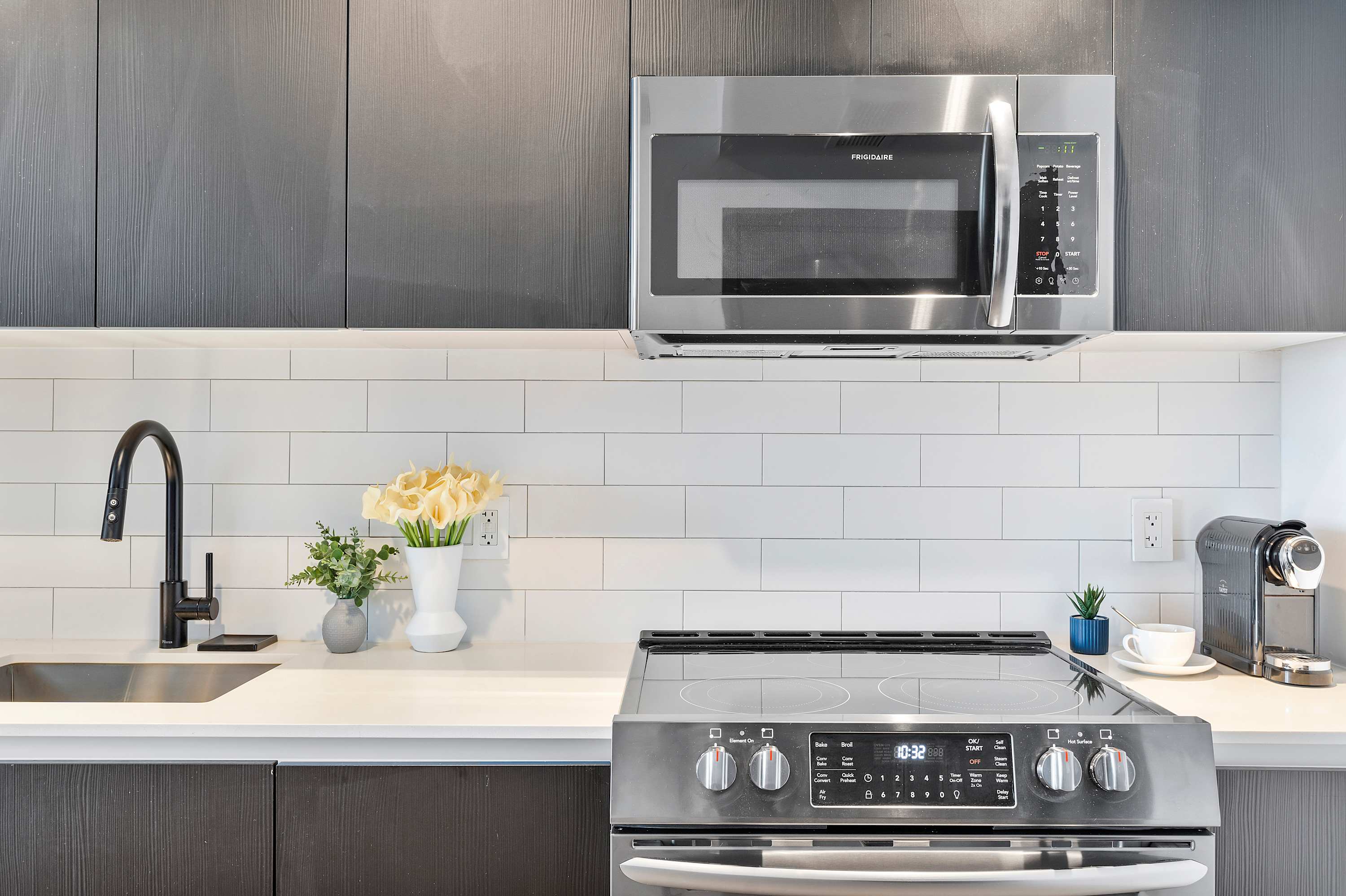 A detailed view of the modern kitchen counter, showcasing a stainless steel microwave above an electric stove, a black faucet over a sink, white subway tile backsplash, and a coffee machine.
