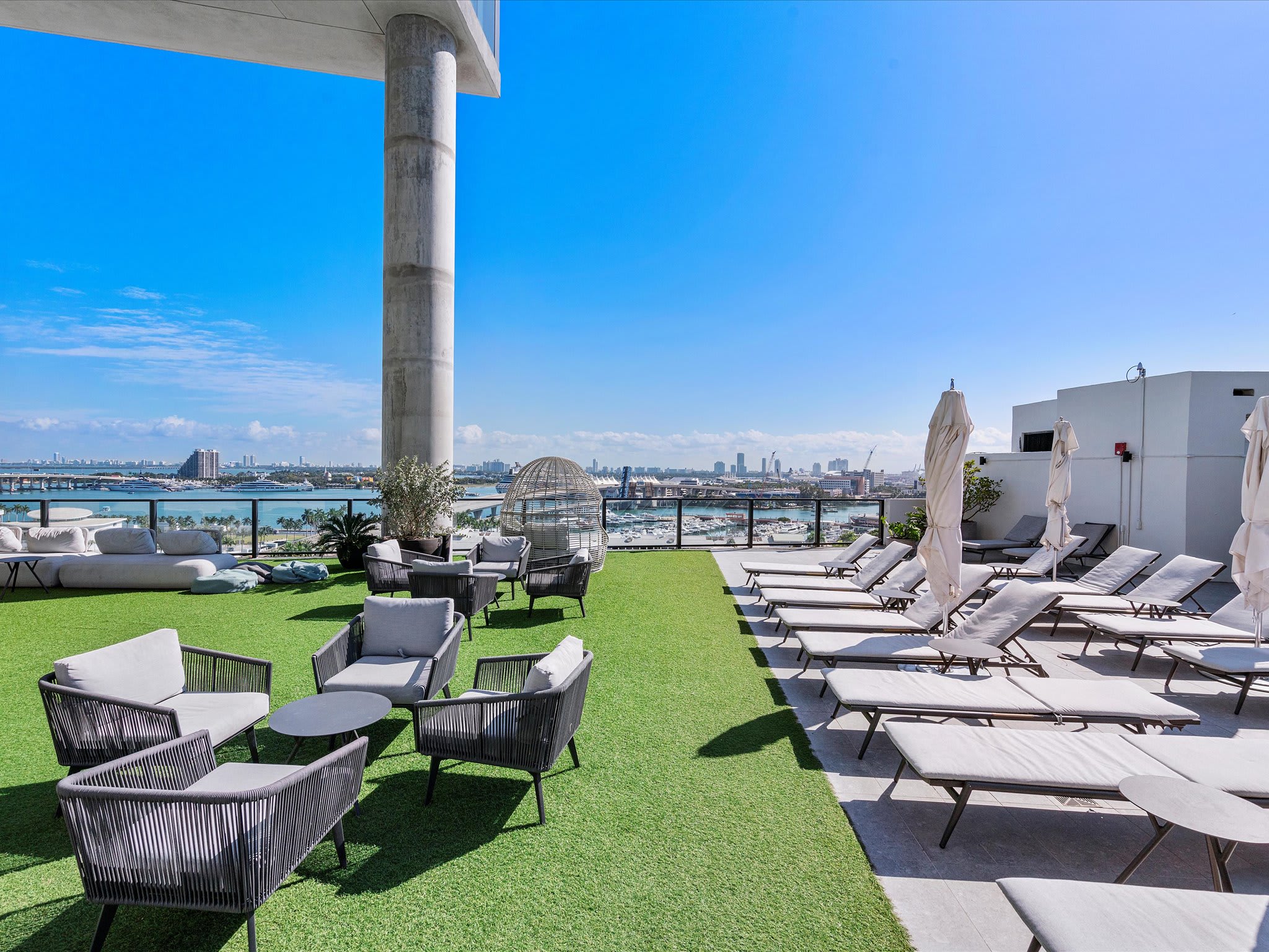 An expansive rooftop terrace featuring a mix of artificial green turf and tiled areas, furnished with comfortable lounge chairs and sun loungers. The space provides stunning views of the city skyline and a busy waterfront with bridges and boats.