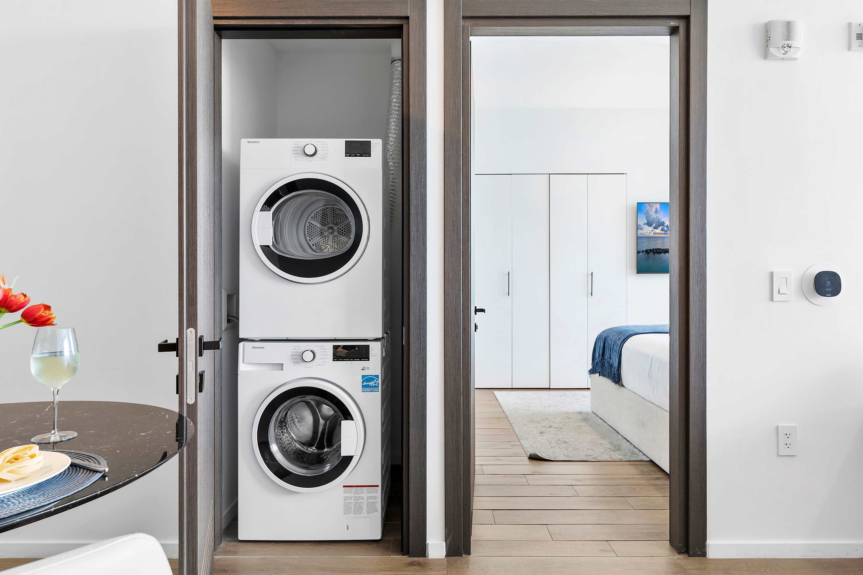 A compact laundry closet housing a stacked white washer and dryer unit, positioned between a dining area and a doorway leading into a bedroom, with wood flooring throughout.