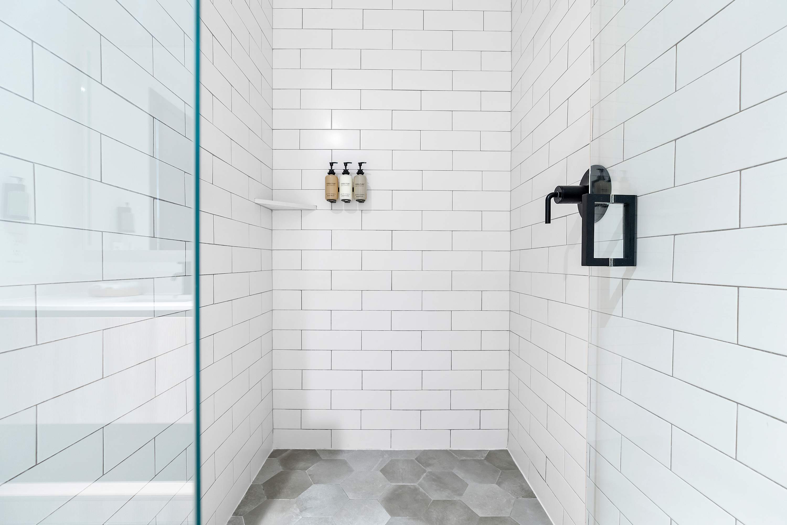 An interior view of the walk-in shower, featuring clean white subway tile walls, a dark square shower head, and a black square handle, with a dark hexagonal tile floor.