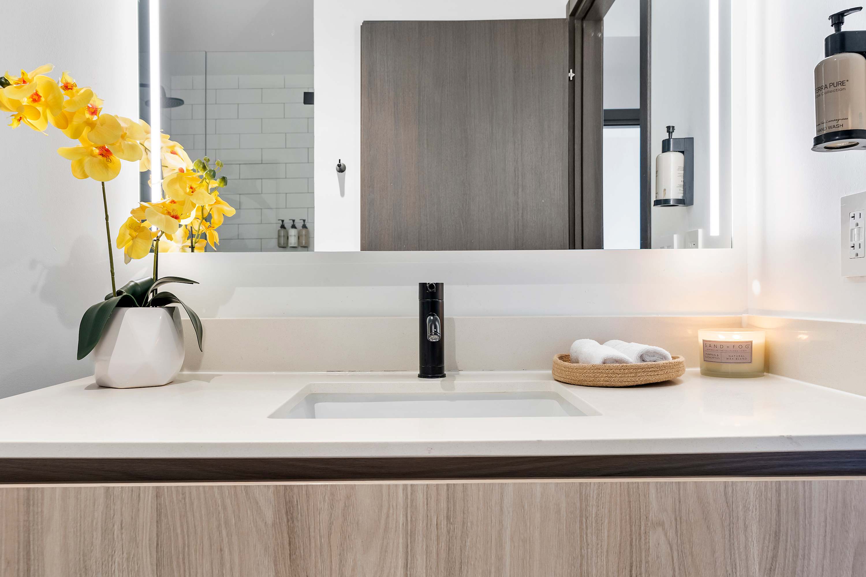 A sleek bathroom vanity with a rectangular undermount sink, a black faucet, and an illuminated mirror, adorned with a vibrant yellow orchid plant and a woven basket of white towels.