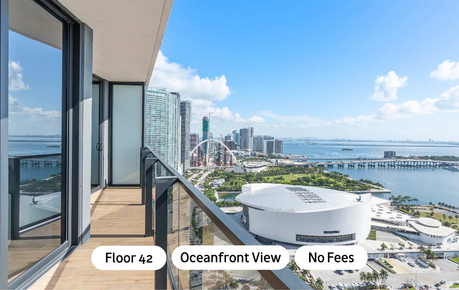 High-rise balcony with glass railing offers panoramic oceanfront and city views, showcasing a stadium and bridges under a clear blue sky. The floor is light wood planking.