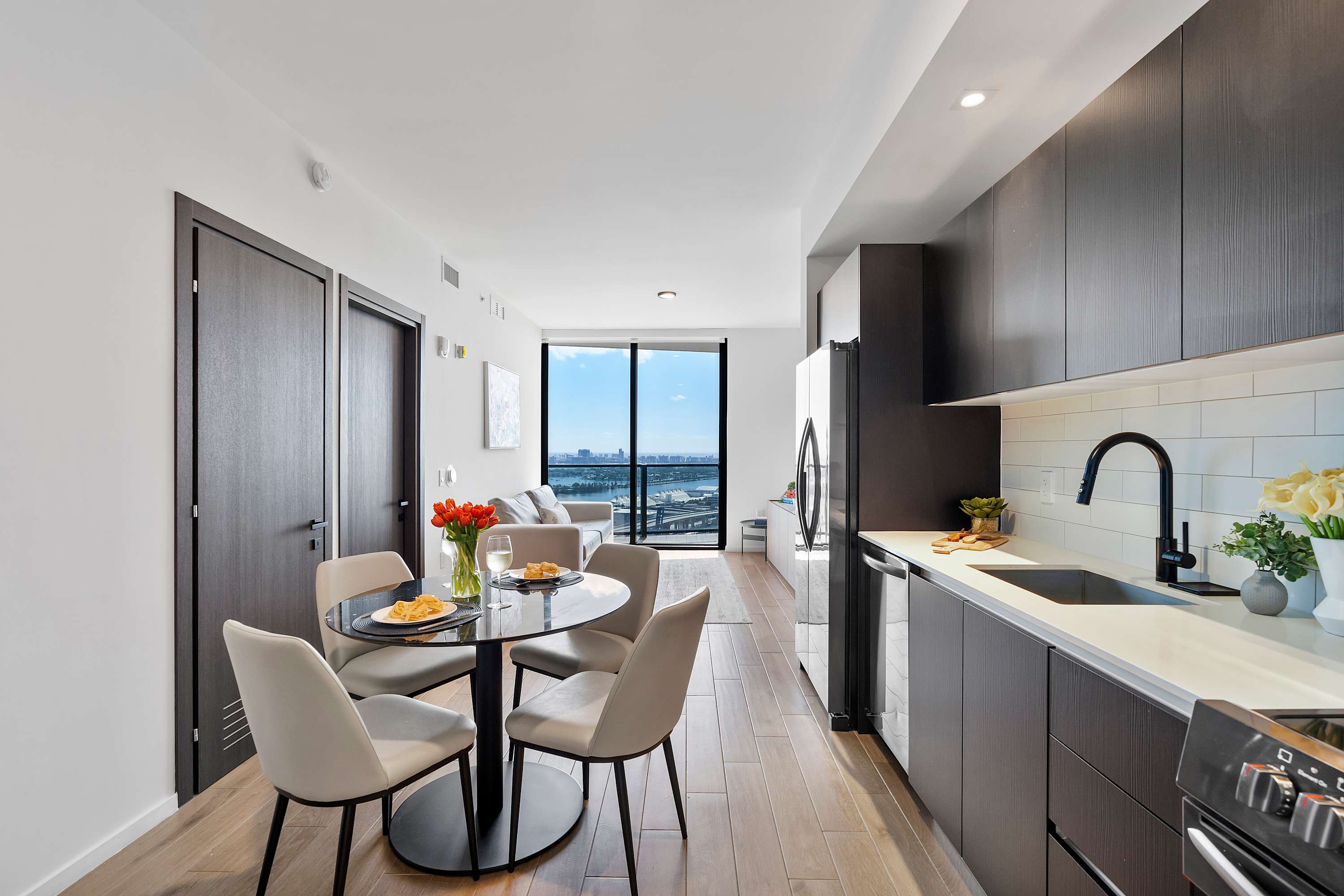 A modern kitchen and dining area featuring dark wood cabinetry, white subway tile backsplash, a black round dining table with four chairs, and a view towards the living area and large windows.