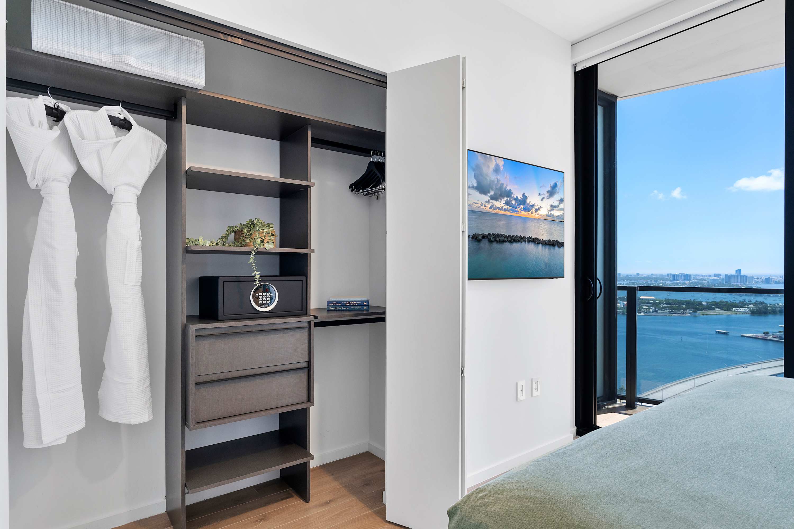 An open closet area within the bedroom, featuring built-in shelving, hanging space with white robes, a small safe, and a glimpse of the wall-mounted TV and city view from the window.