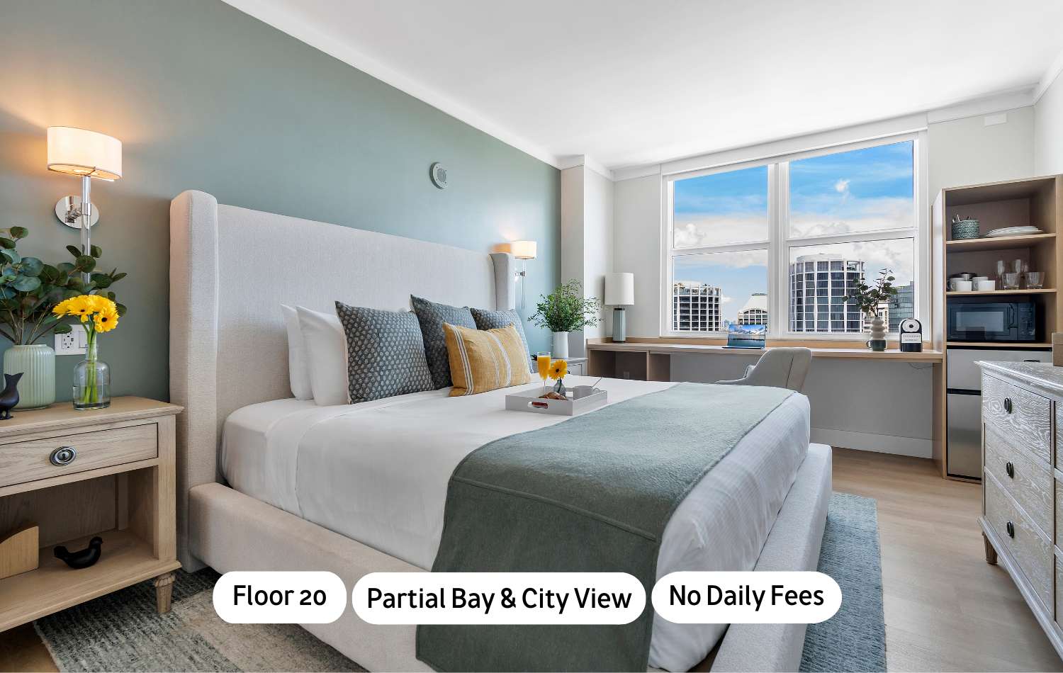A modern bedroom features a light-colored upholstered bed with blue and yellow pillows, a wooden nightstand, and a desk area by a large window offering partial bay and city views. The walls are painted a soothing light green.