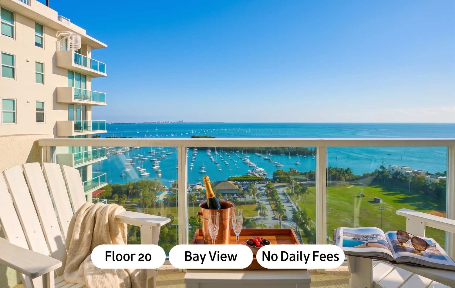 A high-floor balcony offers stunning bay views with numerous boats, featuring two white Adirondack chairs, a small table with champagne and berries, and a building facade on the left.