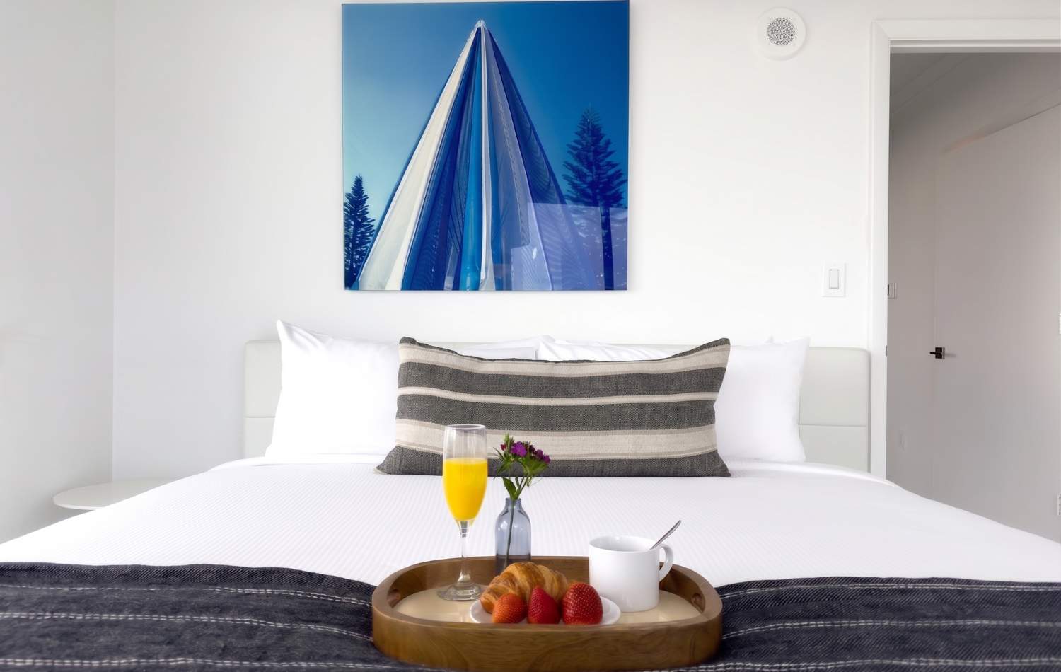 Bright bedroom featuring a comfortable white bed adorned with a striped throw pillow and a breakfast tray holding orange juice, a croissant, and fresh strawberries. A large abstract art piece with blue and white tones hangs prominently above the headboard.