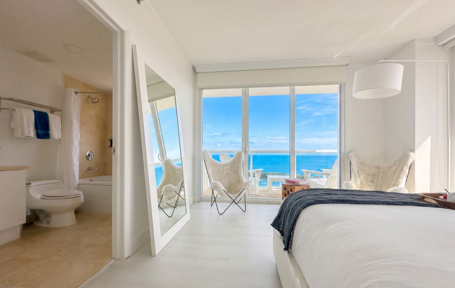 Bright bedroom with a white bed, light wood floors, and floor-to-ceiling windows offering ocean and city views. A full-length mirror reflects the room and the view, with a bathroom entrance visible on the left.