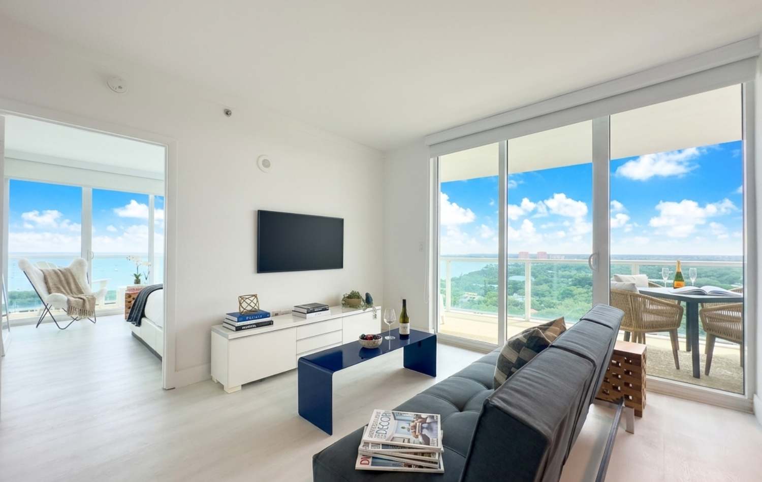 Bright living room with a dark gray sofa, a blue coffee table, and a wall-mounted TV above a white console. Large sliding glass doors lead to a balcony with outdoor seating and a panoramic city and ocean view.