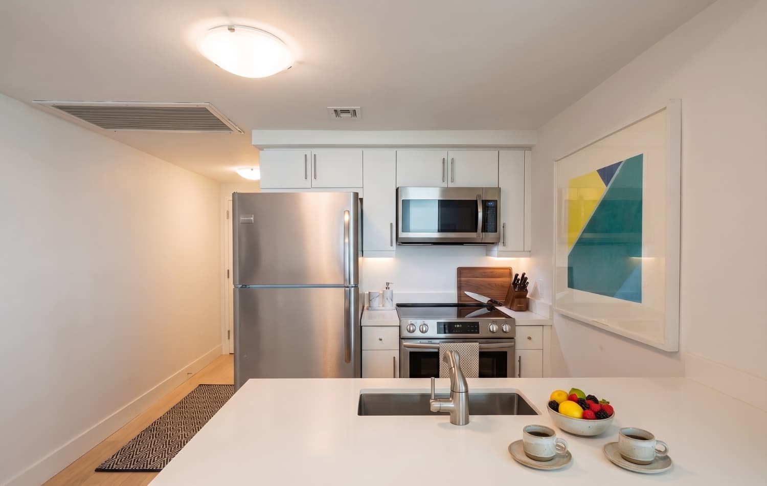 Modern kitchen featuring white flat-panel cabinets, stainless steel appliances including a refrigerator, microwave, and electric range. A white countertop island with a sink and a fruit bowl adds functionality, complemented by light wood flooring and abstract art.