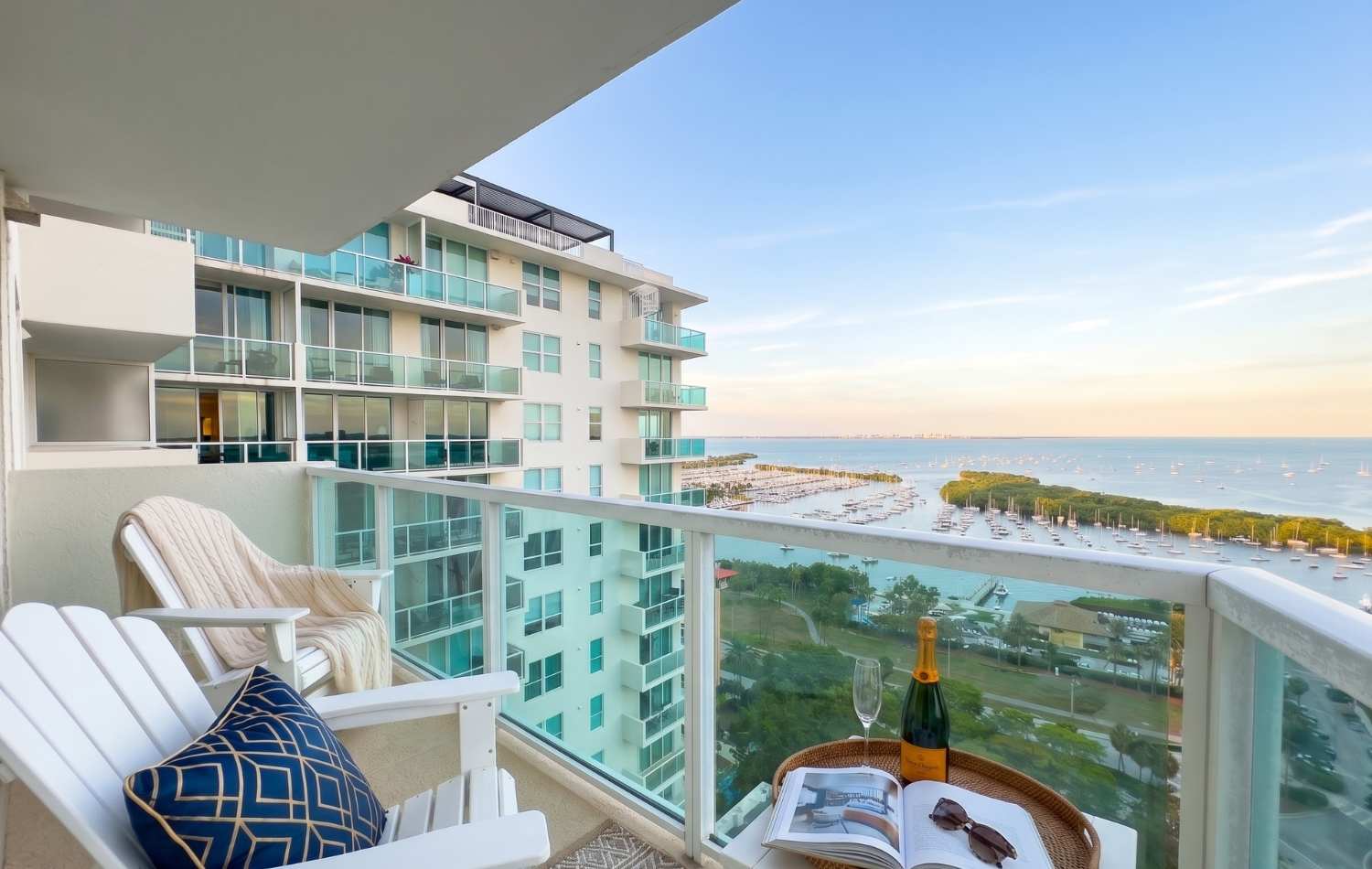 Spacious balcony furnished with two white Adirondack chairs and a small table, offering a stunning panoramic view of a marina filled with boats and the expansive ocean. A bottle of champagne, glasses, and a book are set for relaxation.