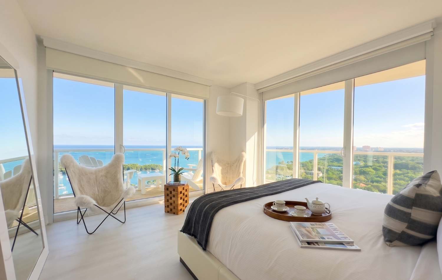 Bright bedroom with a white bed, light wood floors, and floor-to-ceiling windows offering ocean and city views. Two furry butterfly chairs and a small wooden table with an orchid are by the window, with a breakfast tray on the bed.
