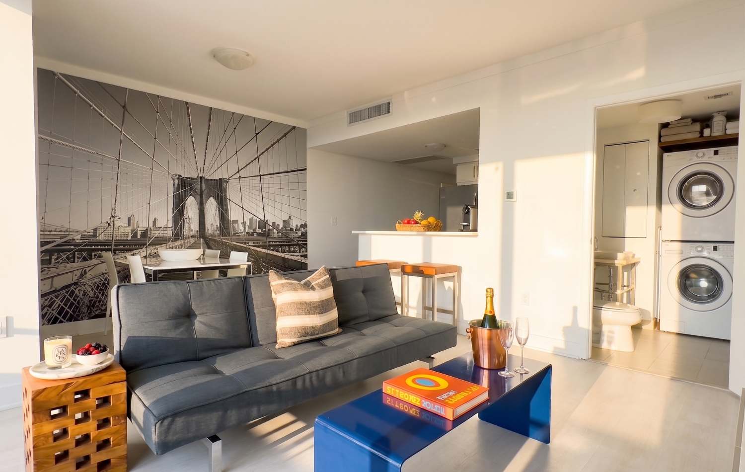 Contemporary living room featuring a dark gray futon sofa, a vibrant blue coffee table, and a striking black and white mural of the Brooklyn Bridge on the accent wall. A small bar area with stools is visible in the background, connecting to the kitchen.