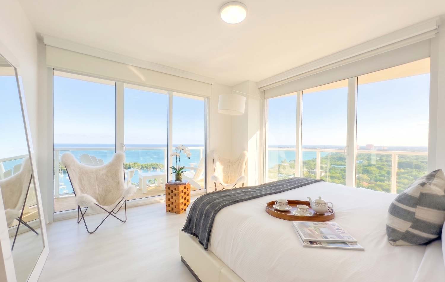 Bright bedroom with a white bed, light wood floors, and floor-to-ceiling windows offering ocean and city views. Two furry butterfly chairs and a small wooden table with an orchid are by the window.