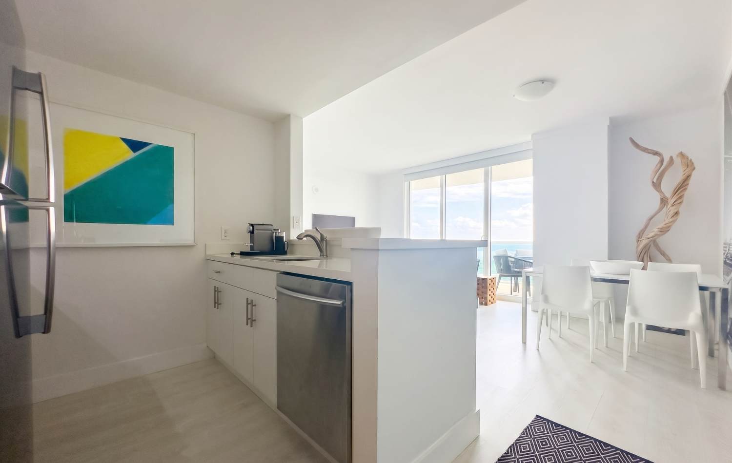 Modern kitchen with white cabinets, a stainless steel dishwasher, and a white countertop peninsula that separates the kitchen from the dining area. A colorful abstract art piece hangs on the wall, and a coffee maker is visible on the counter.