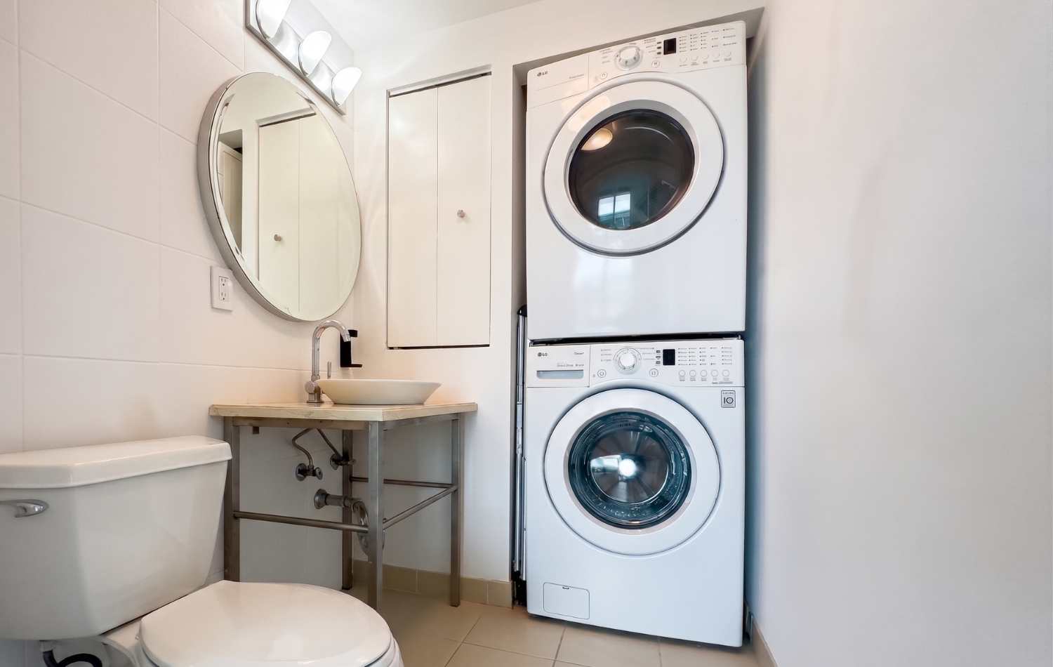 Compact laundry area featuring a stacked white LG washer and dryer unit, positioned next to a small bathroom with a toilet and a modern vessel sink on a metal stand. The space is bright with white walls and tiled accents.