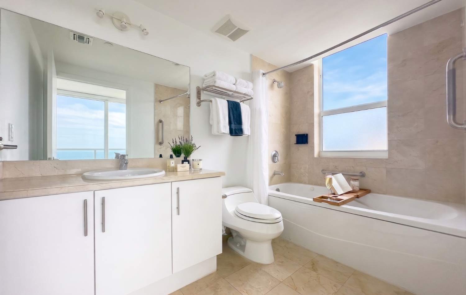 Modern bathroom with light beige tiled floors and walls, featuring a white vanity with a single sink, a toilet, and a bathtub with a shower combo and a window offering an ocean view.