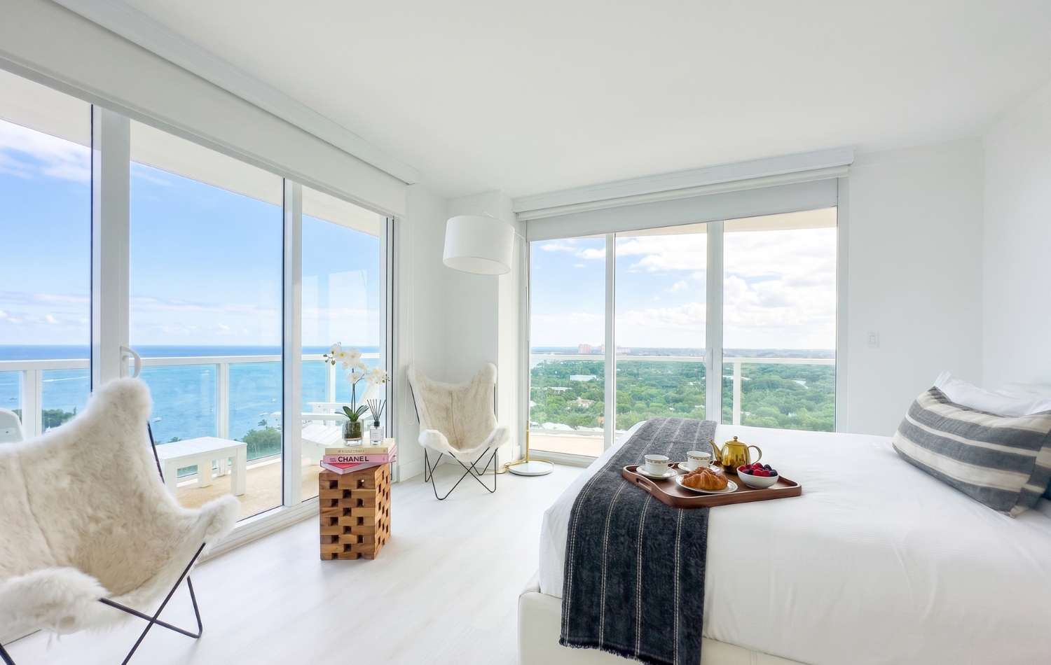 Bright bedroom with a white bed, light wood floors, and floor-to-ceiling windows offering ocean and city views. Two furry butterfly chairs and a small wooden table with an orchid are by the window, with a breakfast tray on the bed.