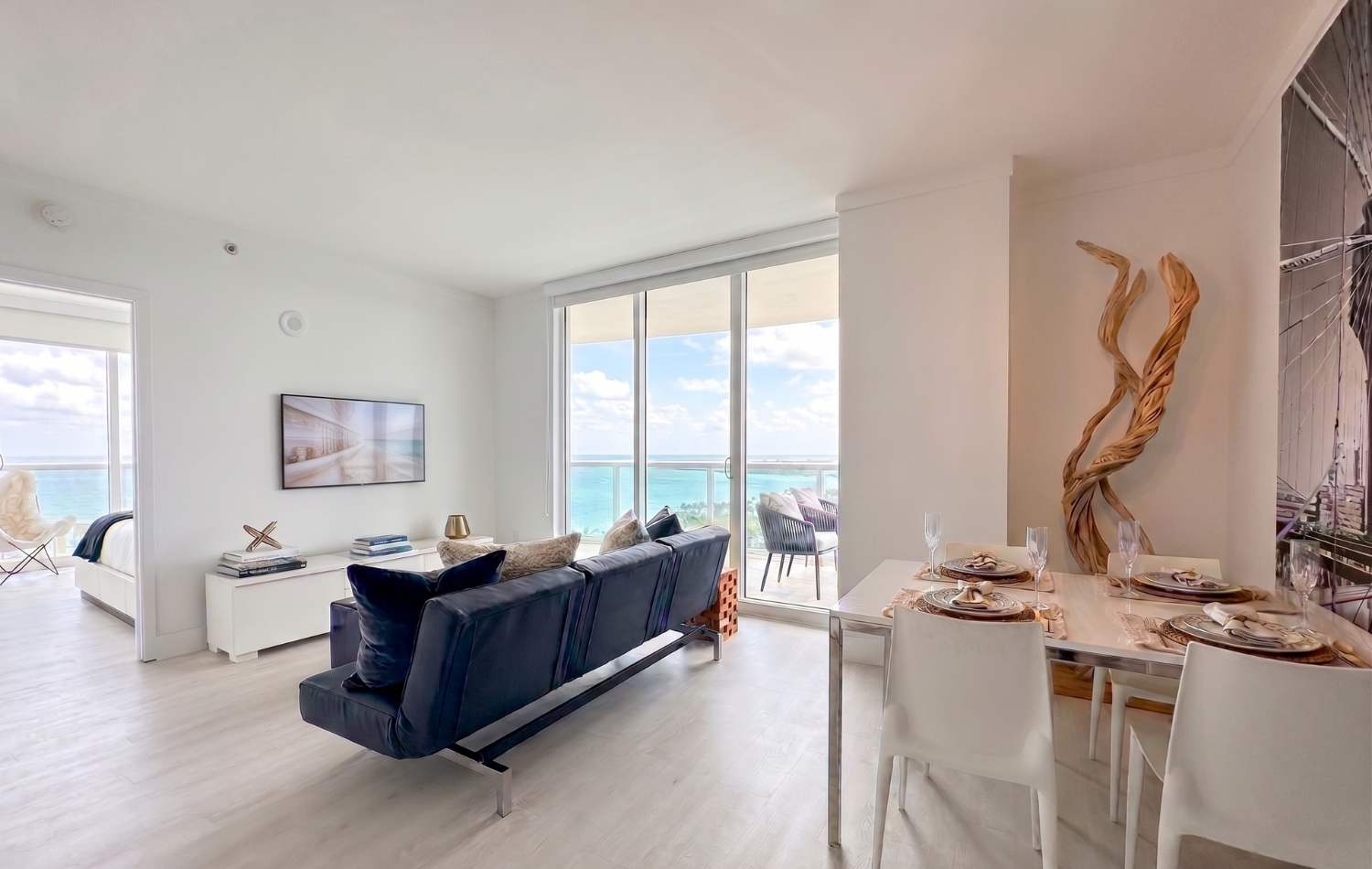 Open-concept living and dining area featuring a dark blue sofa, a white dining table with four chairs, and a large sliding glass door offering expansive ocean views. A unique twisted wood sculpture stands as a decorative element in the corner.