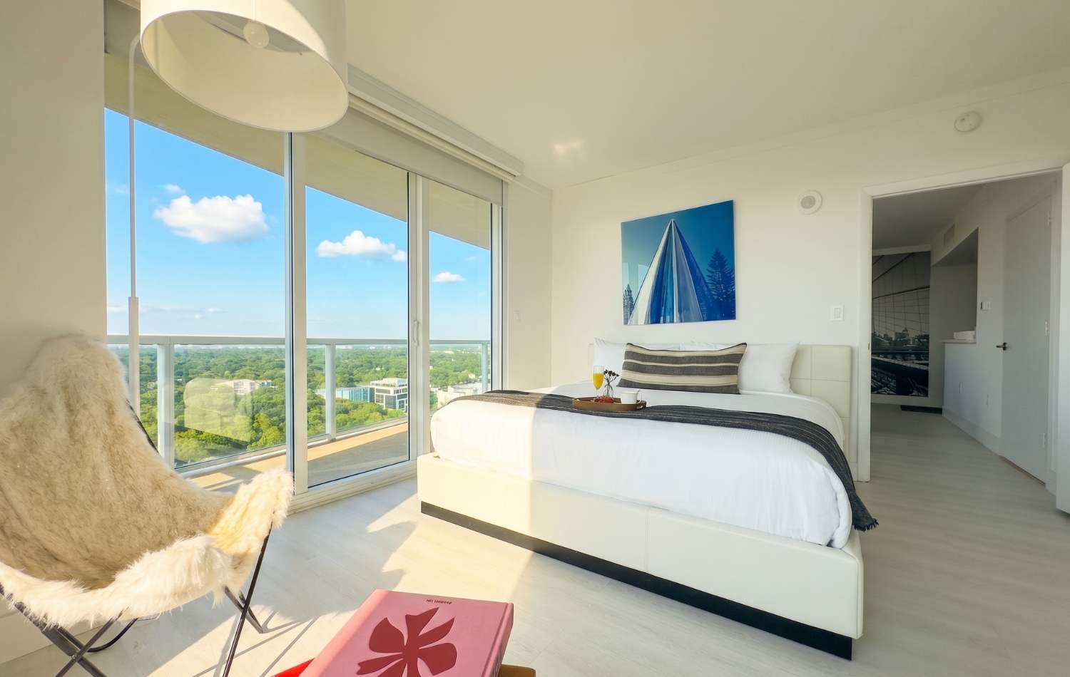 Modern bedroom with a white bed, light wood floors, and large sliding glass doors leading to a balcony with a city view. A blue and white abstract art piece hangs above the bed, and a furry butterfly chair is in the foreground.