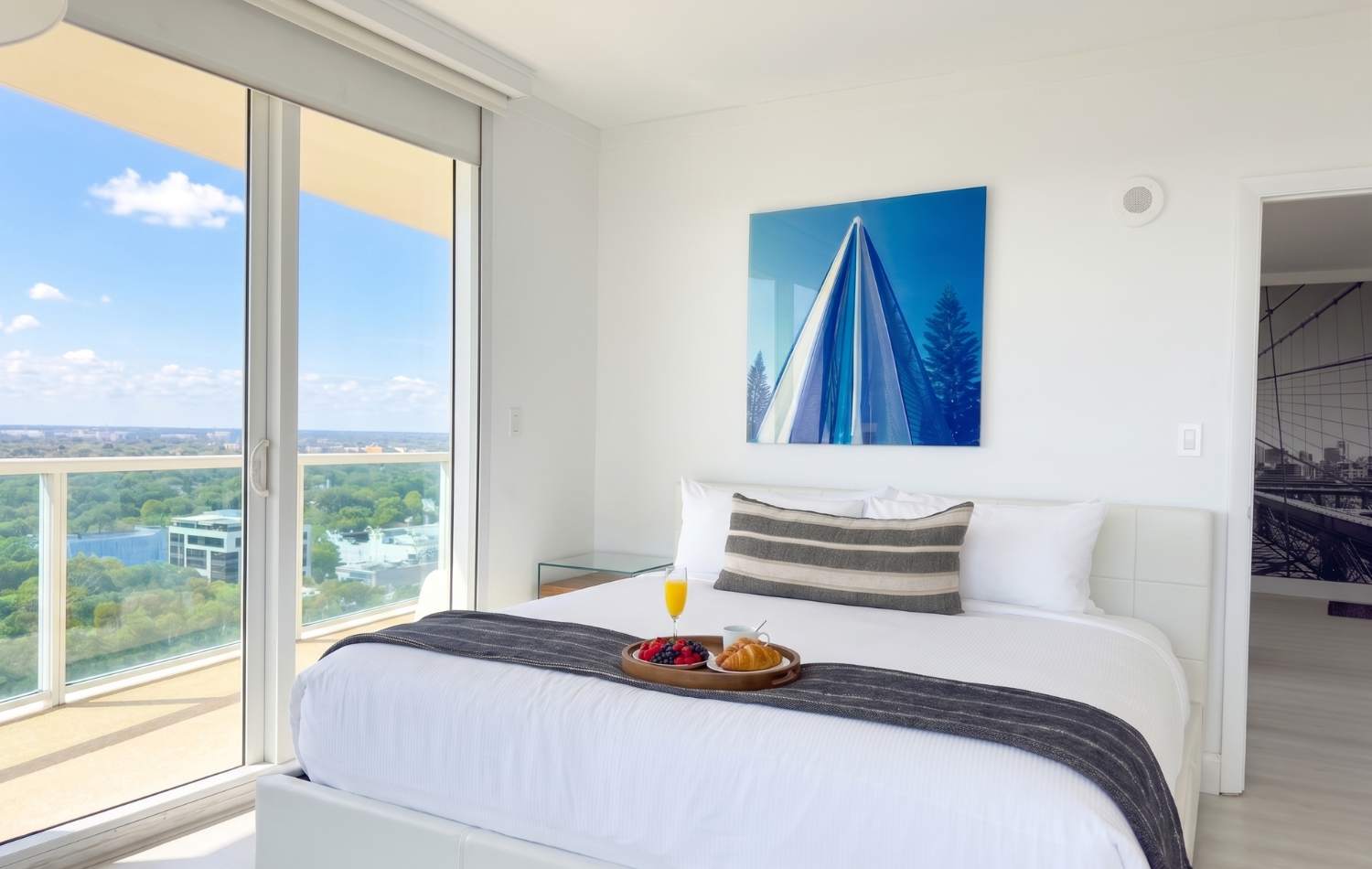 Modern bedroom with a white bed, light wood floors, and large sliding glass doors leading to a balcony with a city view. A blue and white abstract art piece hangs above the bed, and a breakfast tray is on the bed.