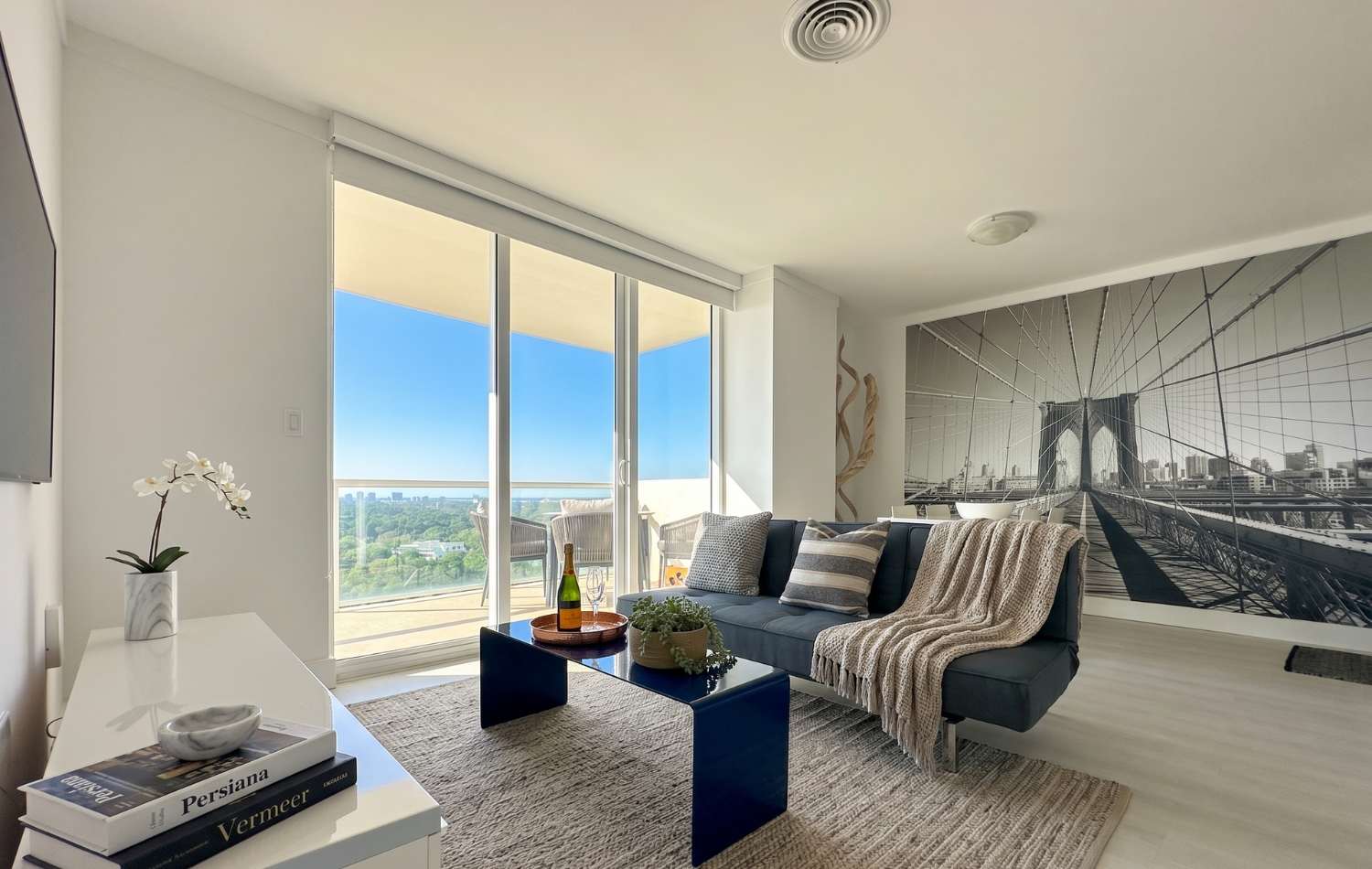 Bright living room with a dark gray futon sofa, a blue coffee table, and a large sliding glass door leading to a balcony with city views. A prominent black and white Brooklyn Bridge mural covers an accent wall, adding an urban touch.