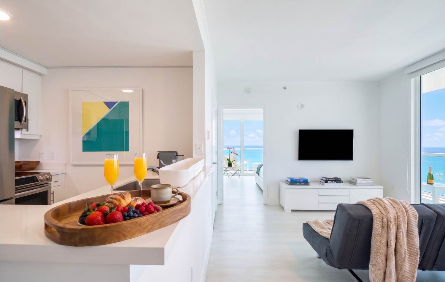 Open-concept living area with a white kitchen counter in the foreground, featuring a breakfast tray with juice and pastries. Beyond, a white living room with a wall-mounted TV and a glimpse of the ocean through a doorway.