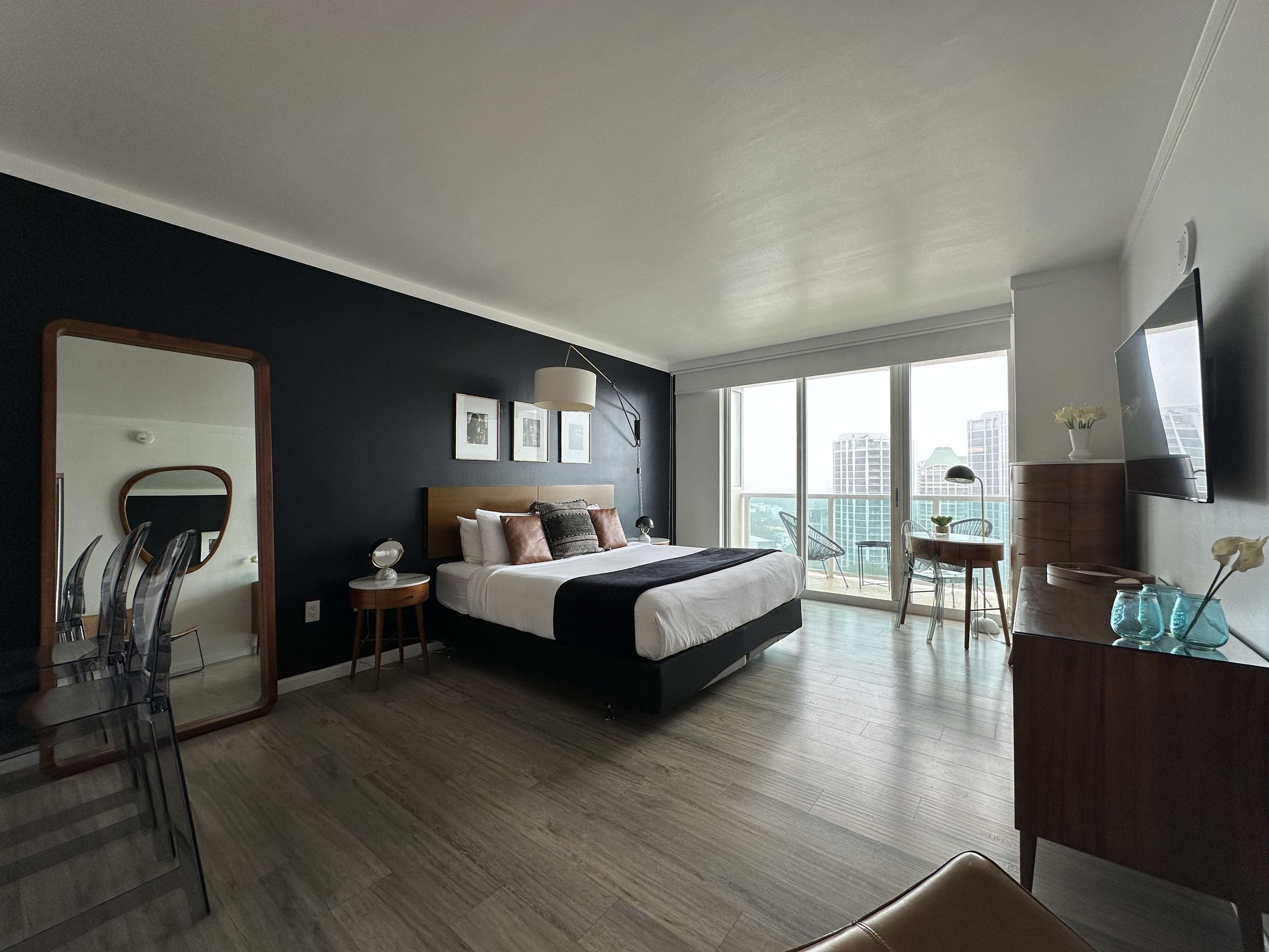 Expansive view of a modern studio apartment, showcasing a king bed, a small desk by large windows offering city views, and the integrated kitchen area in the background. Light wood-look flooring throughout.