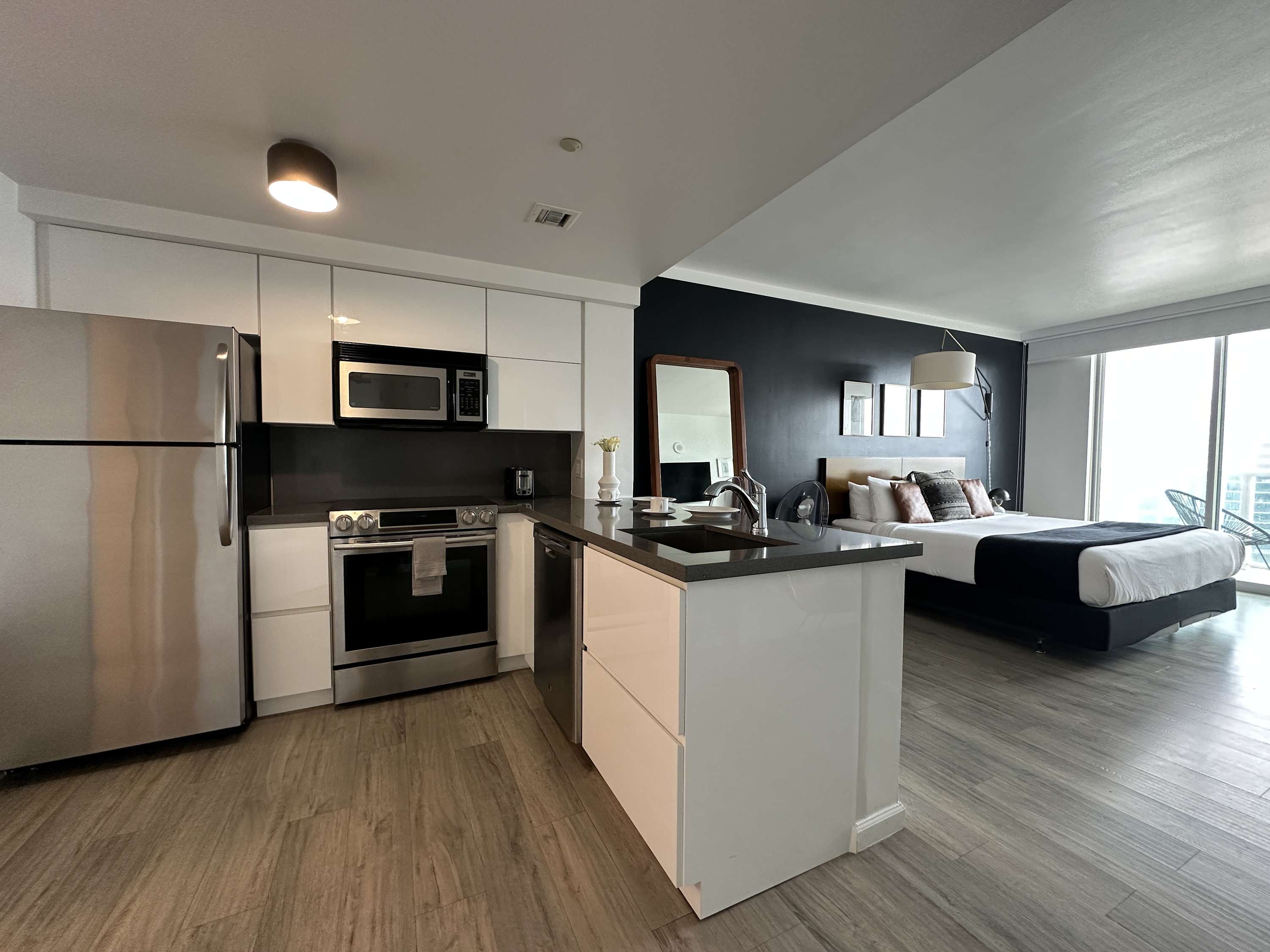 Contemporary kitchen area within a studio apartment, equipped with white cabinetry, stainless steel appliances including a refrigerator and oven, and a dark countertop with two clear bar stools.