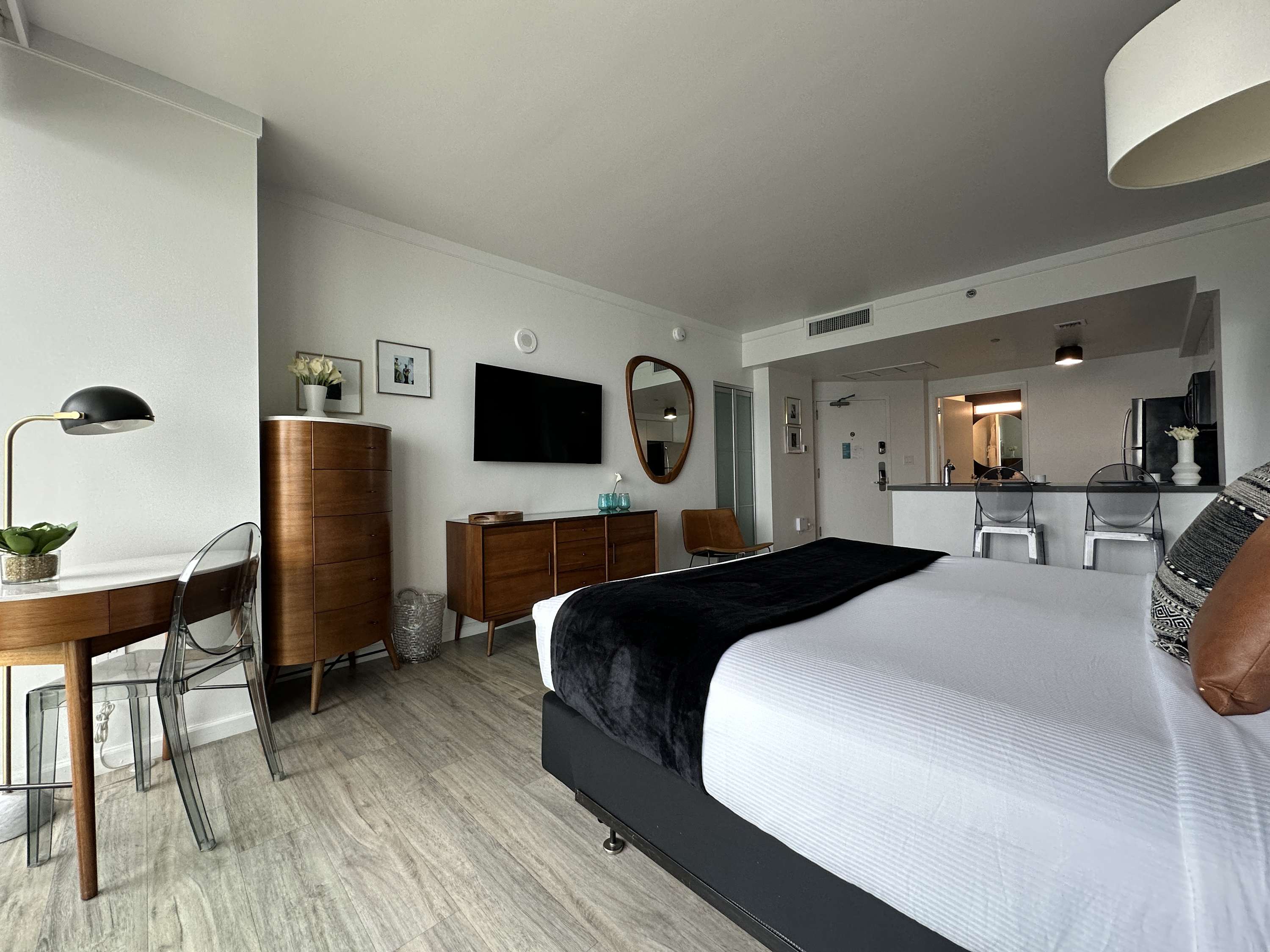 Bright studio apartment interior featuring a neatly made bed, a wooden desk with a clear chair, a dresser with a television, and a large full-length mirror. The room has light wood-look flooring.