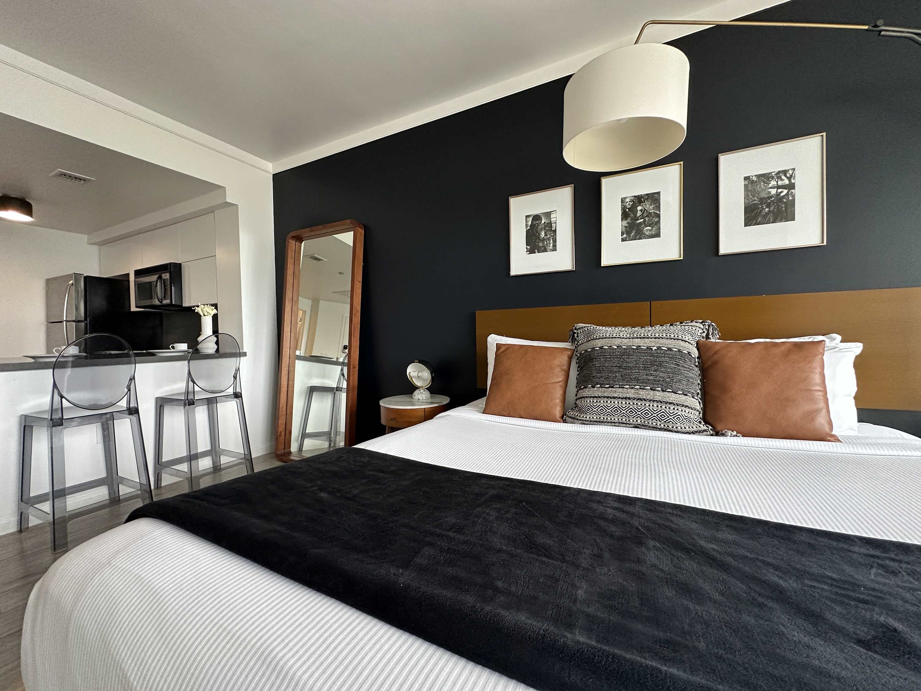 Cozy bedroom area with a king-sized bed, a wooden headboard, and decorative pillows against a dark accent wall adorned with three framed pictures. The kitchen bar is visible in the background.