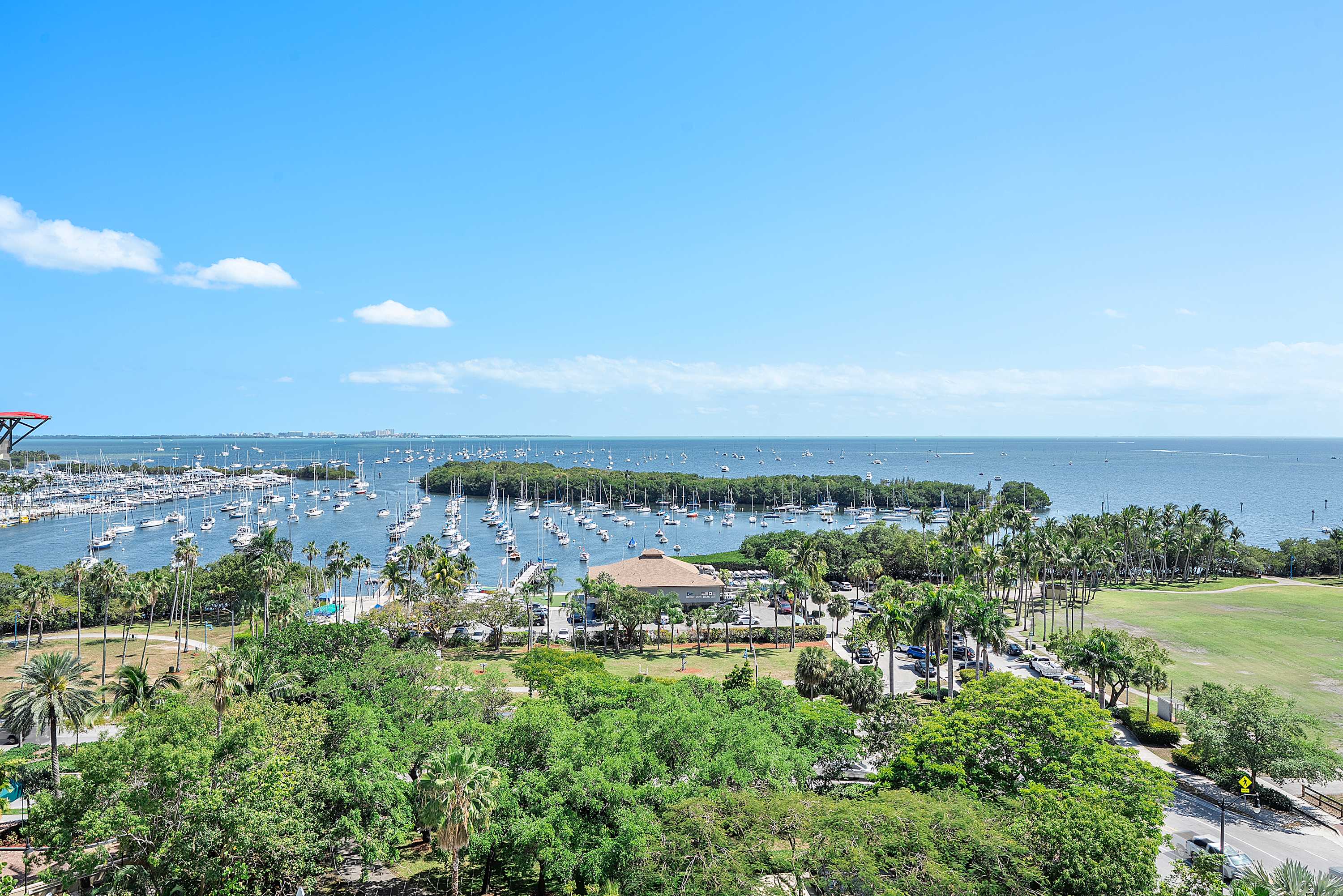 Expansive aerial view showcasing a vibrant marina teeming with numerous sailboats and yachts, surrounded by lush green parks, palm trees, and the vast blue waters of the bay extending to the horizon.