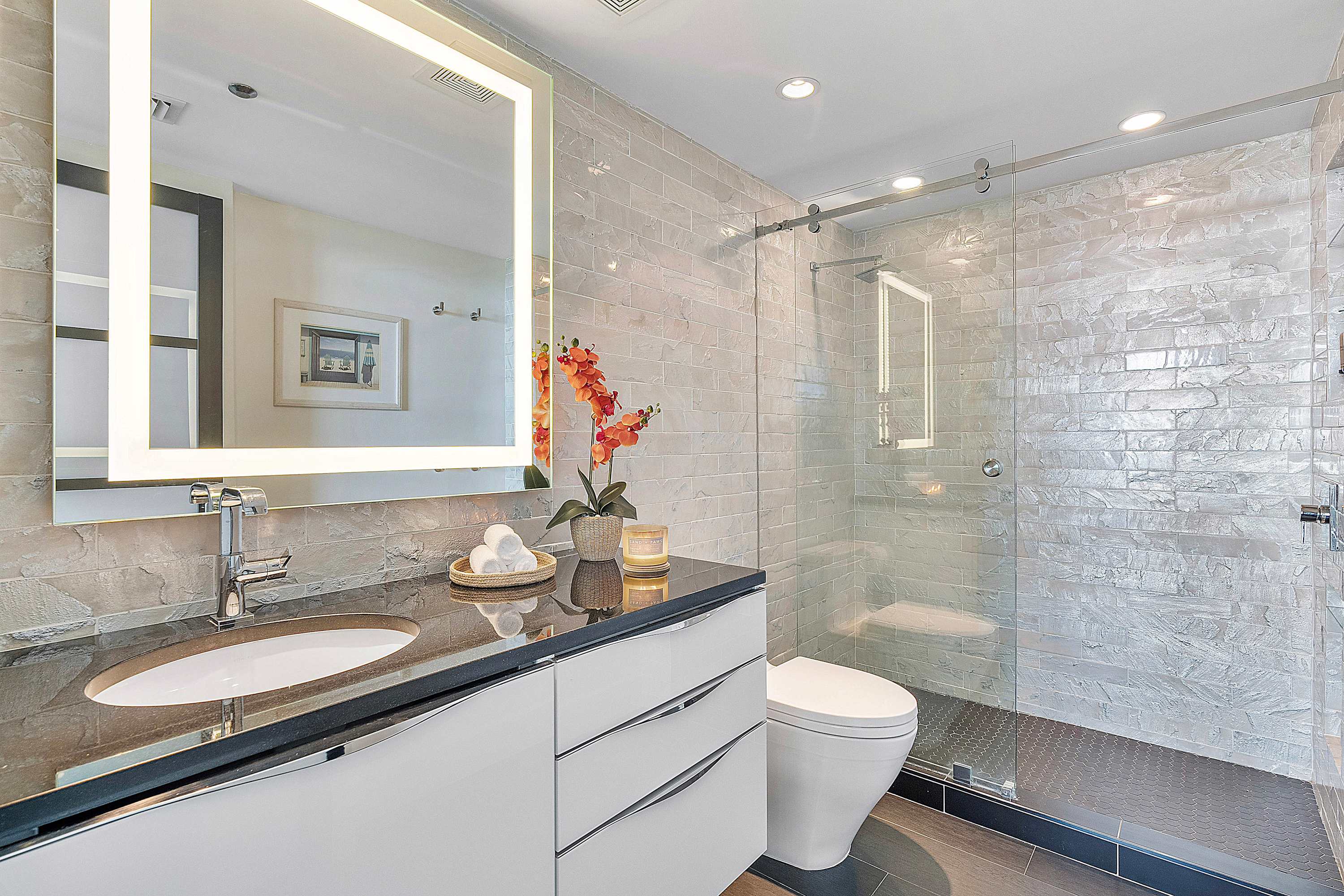 Modern bathroom featuring a dark vanity with a single sink and white drawers, a large illuminated mirror, and a glass-enclosed walk-in shower with textured grey subway tiles.