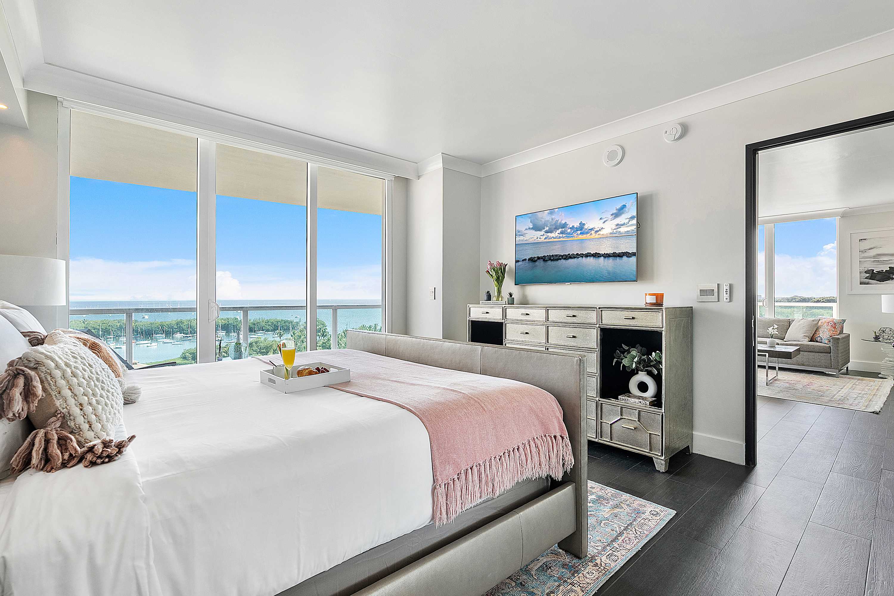 A bright and modern bedroom featuring a large bed with white and pink bedding, a flat-screen TV on a stylish dresser, and expansive floor-to-ceiling windows offering panoramic ocean and marina views.