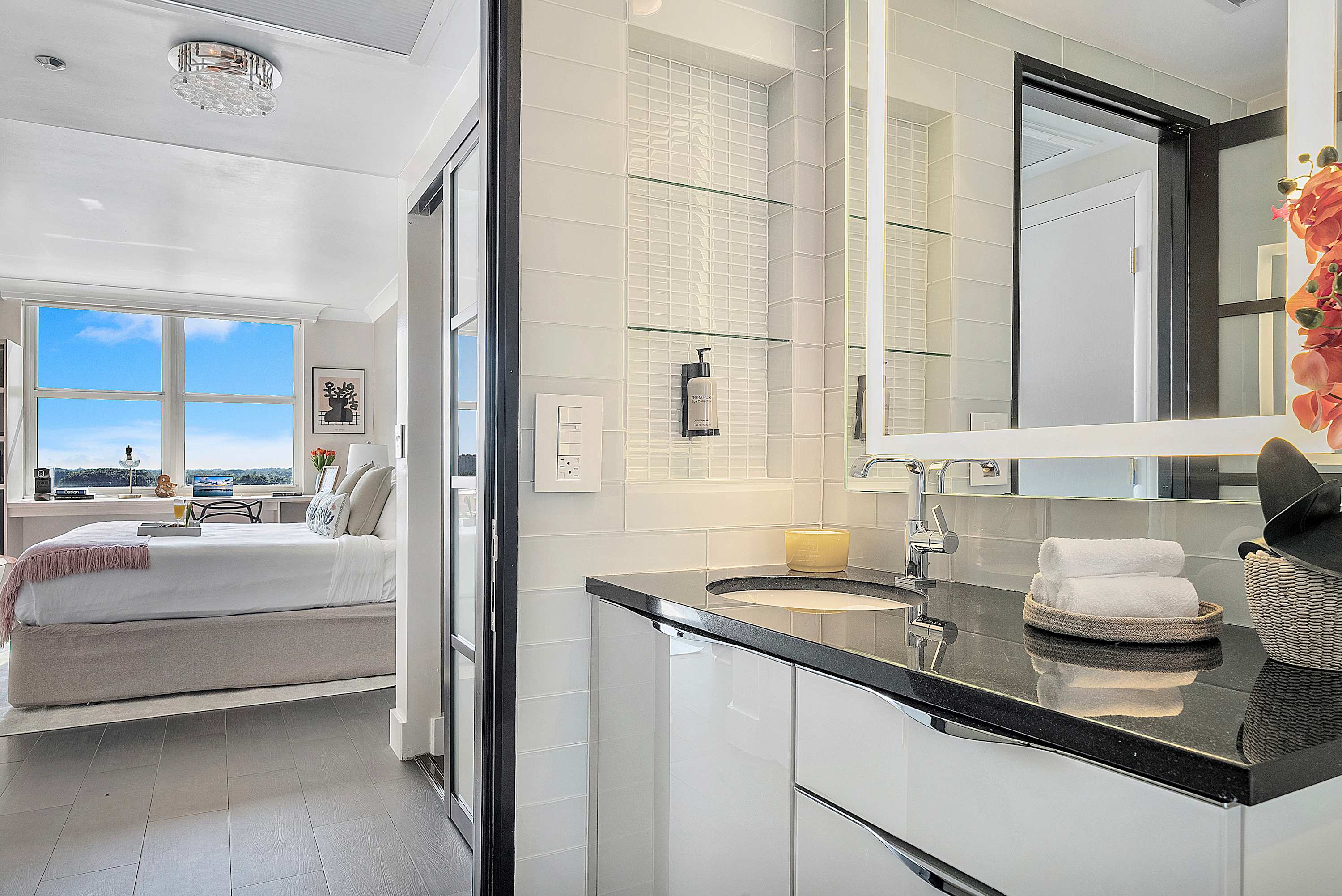 Close-up view of the modern bathroom vanity with a dark countertop, white cabinets, and a large illuminated mirror, offering a glimpse into the adjacent bedroom through a stylish dark-framed doorway.