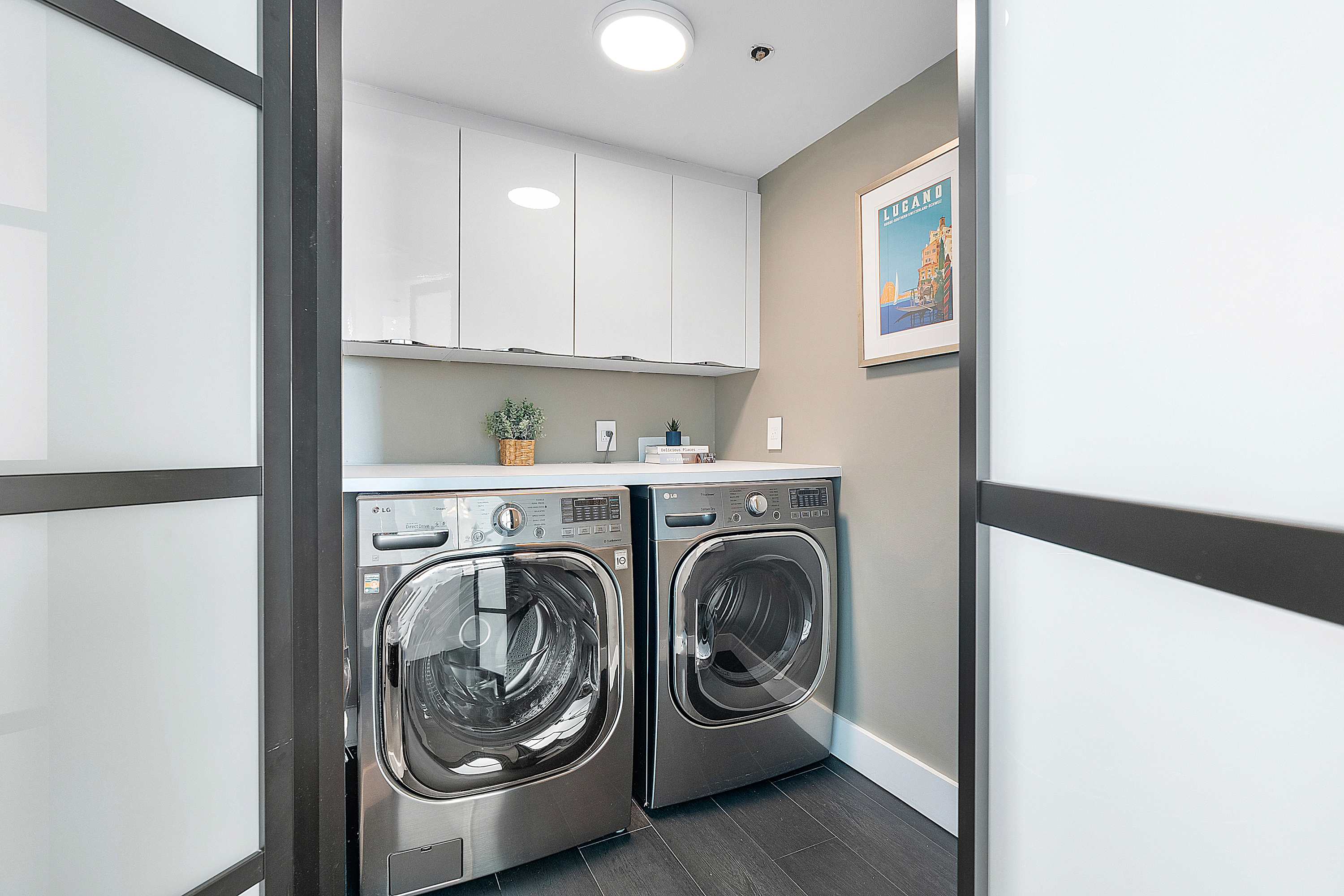 A dedicated laundry room featuring a stainless steel front-loading washer and dryer, white upper cabinets for storage, and a light-colored countertop, with dark tiled flooring.