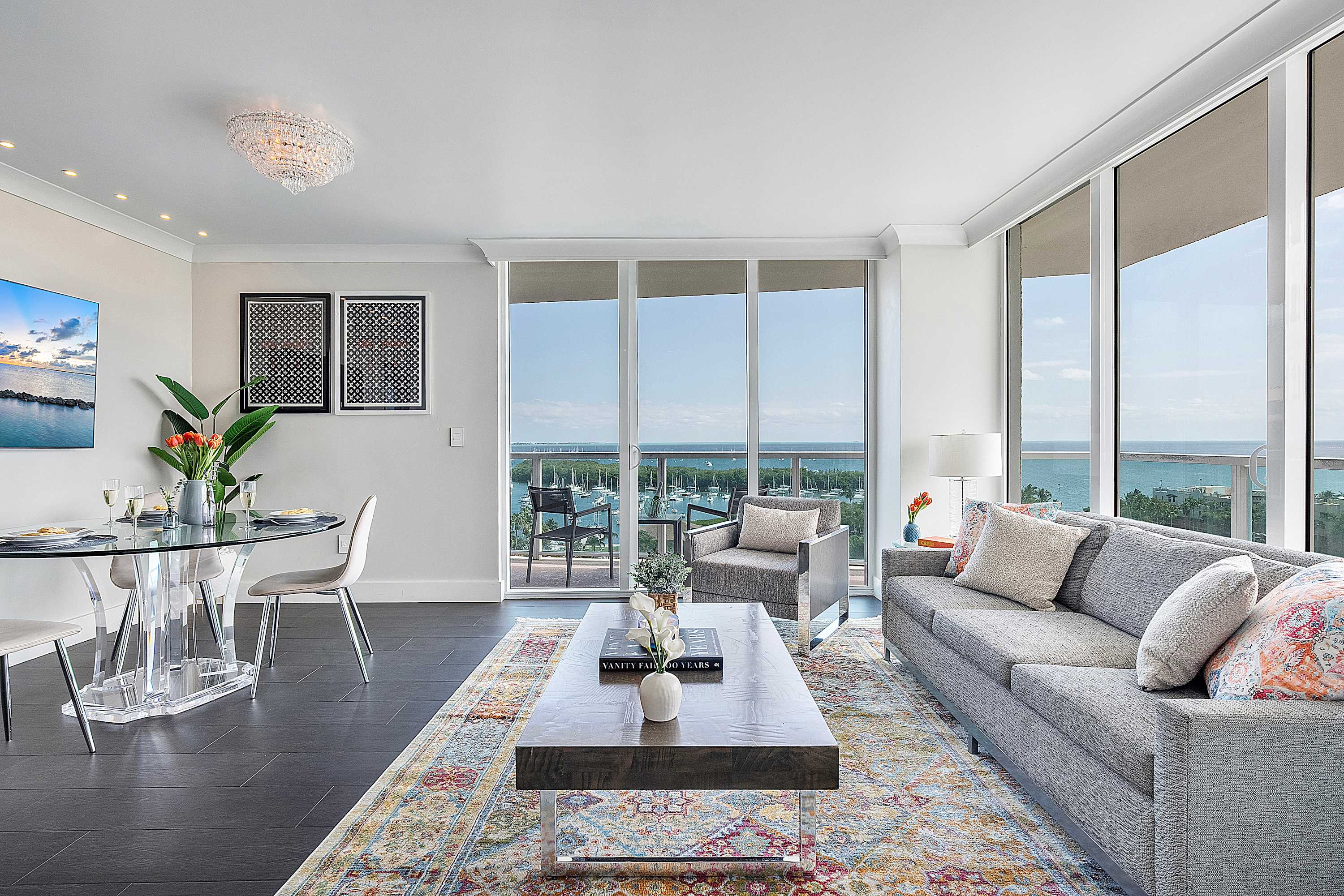 An elegant open-concept living and dining area with dark wood floors, a patterned area rug, a grey sofa, and a glass dining table, all framed by floor-to-ceiling windows overlooking the ocean and a balcony.