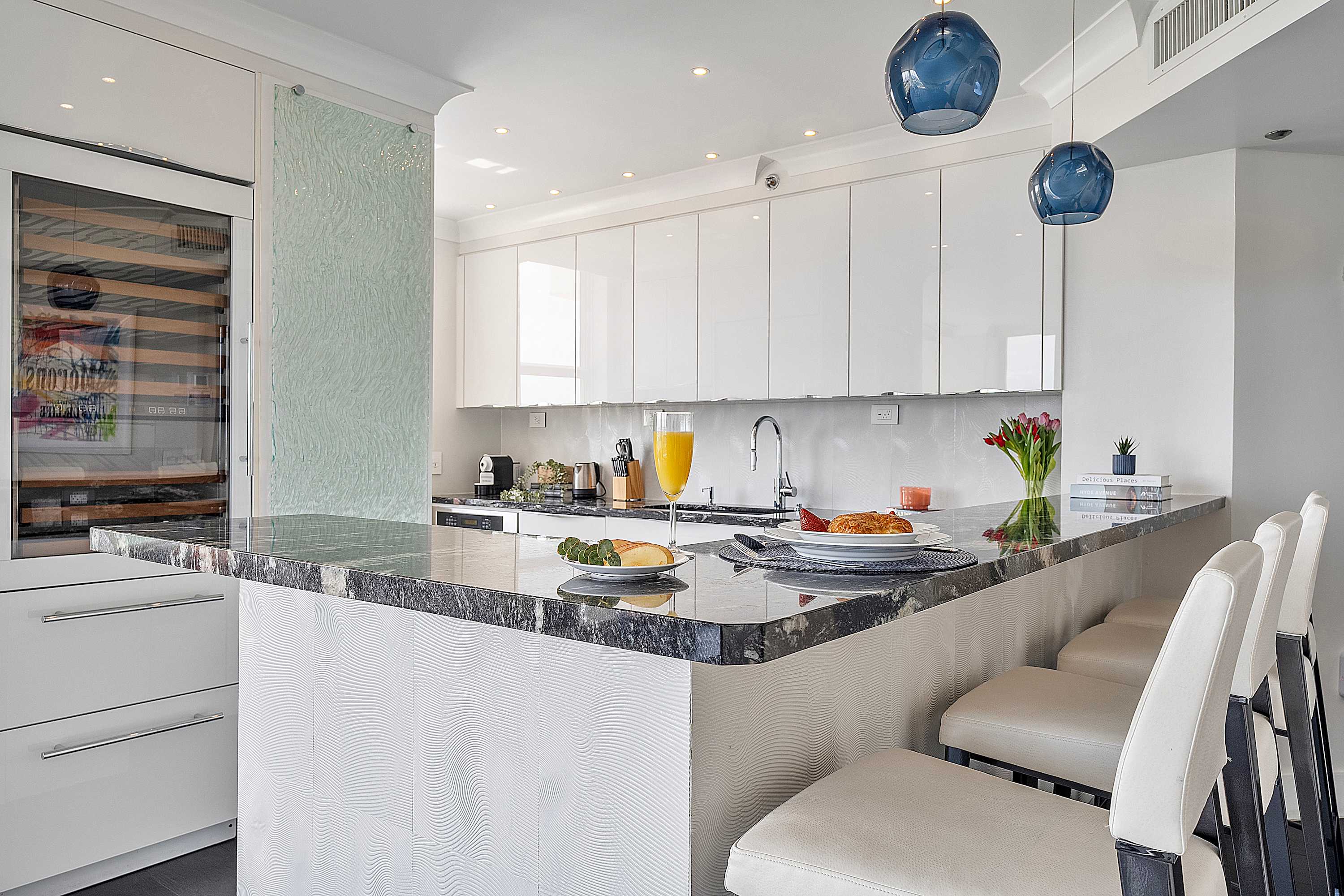 Sleek, modern kitchen featuring white glossy flat-panel cabinetry, a large dark granite countertop island with bar seating, and two unique blue glass pendant lights. A built-in wine cooler is visible on the left.