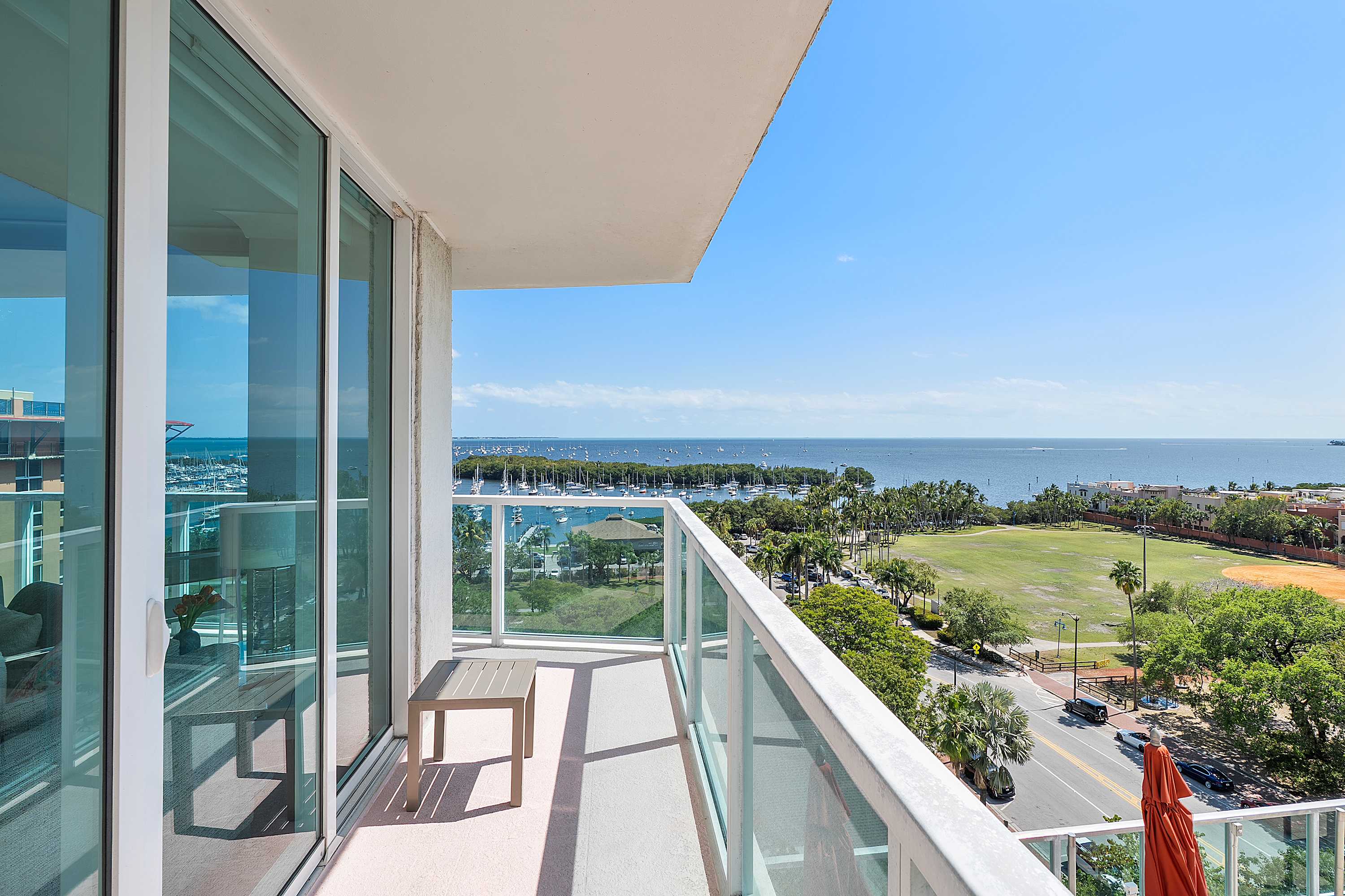 A private balcony with clear glass railings and a small outdoor table, offering a breathtaking panoramic view of the ocean, marina, and surrounding cityscape under a clear blue sky.
