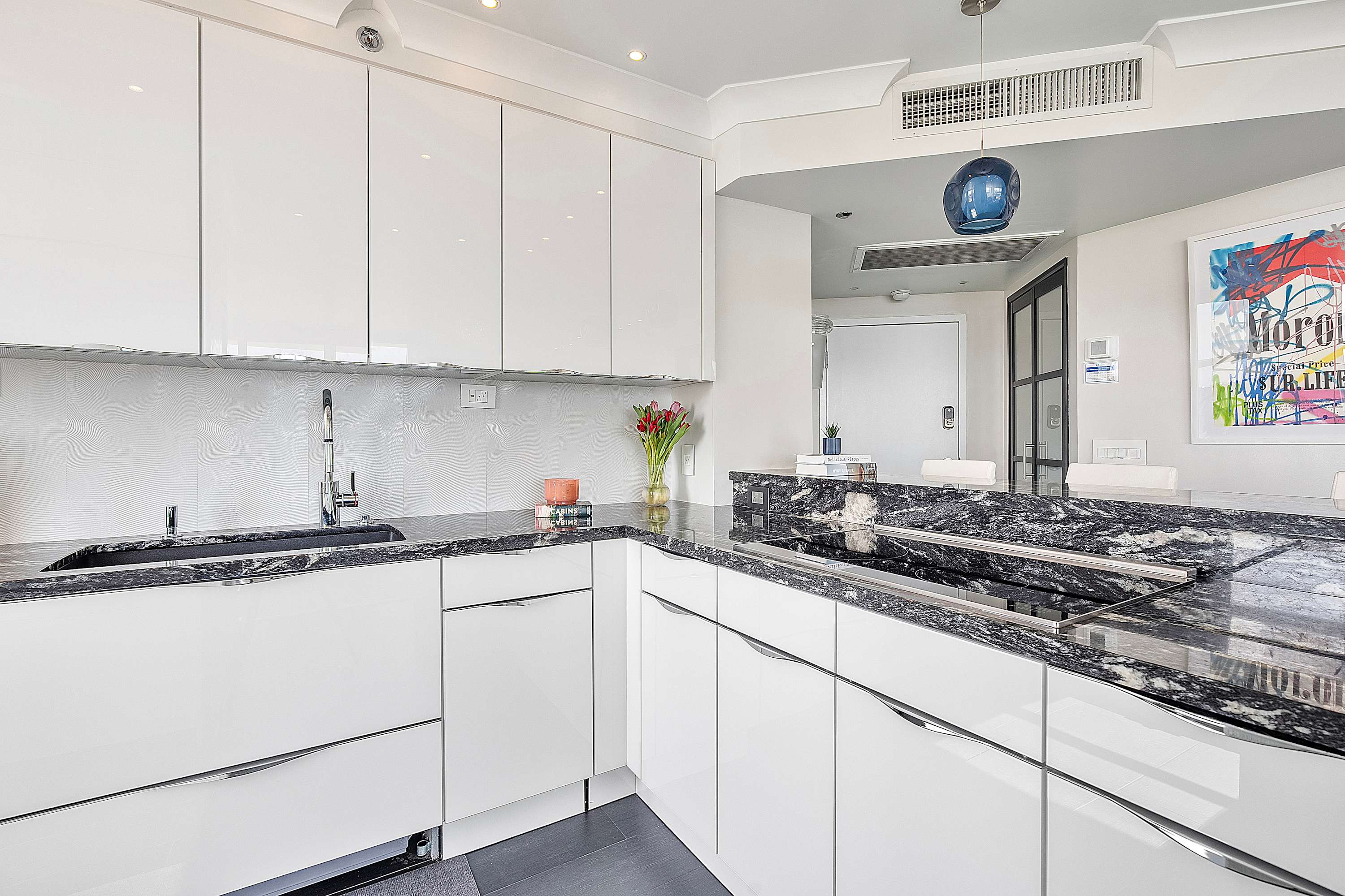A sleek, modern kitchen with white high-gloss cabinetry, dark granite countertops, an integrated cooktop, and a stainless steel sink, illuminated by recessed lighting and a stylish pendant light.