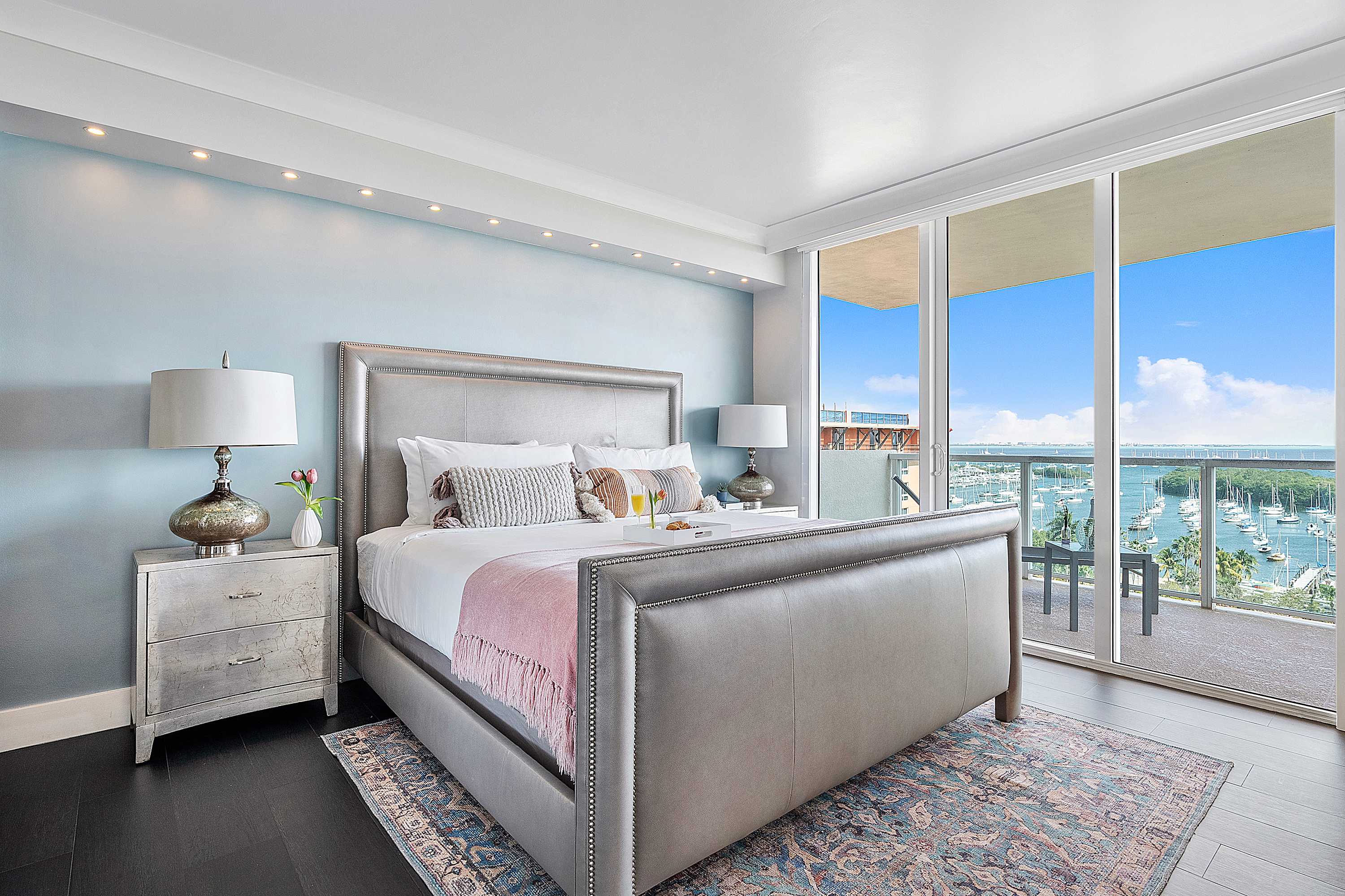Luxurious bedroom with a king-sized bed featuring a grand upholstered headboard, a light blue accent wall, and floor-to-ceiling glass doors leading to a private balcony with stunning bay views.