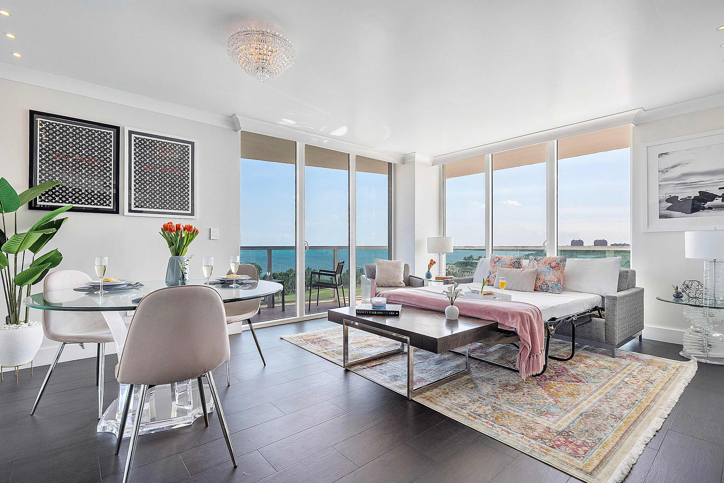 A spacious living area featuring a pull-out sofa bed with pink throw, a dark wood coffee table, and a colorful area rug, complemented by a glass dining table and expansive ocean views through large windows.