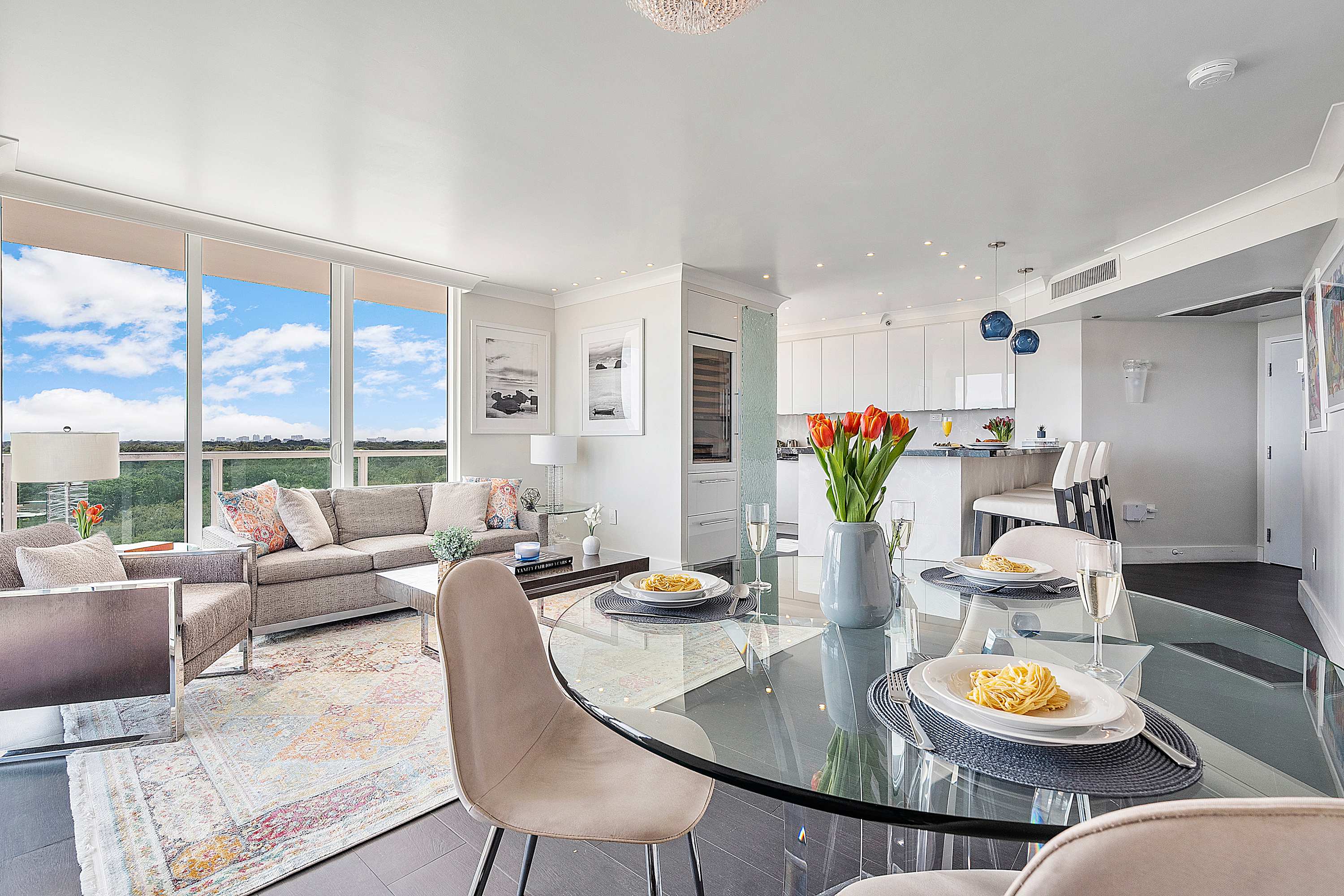 A bright and airy living and dining space with a grey sofa, a glass dining table, and a view into a modern white kitchen, featuring expansive windows that offer stunning ocean vistas and natural light.