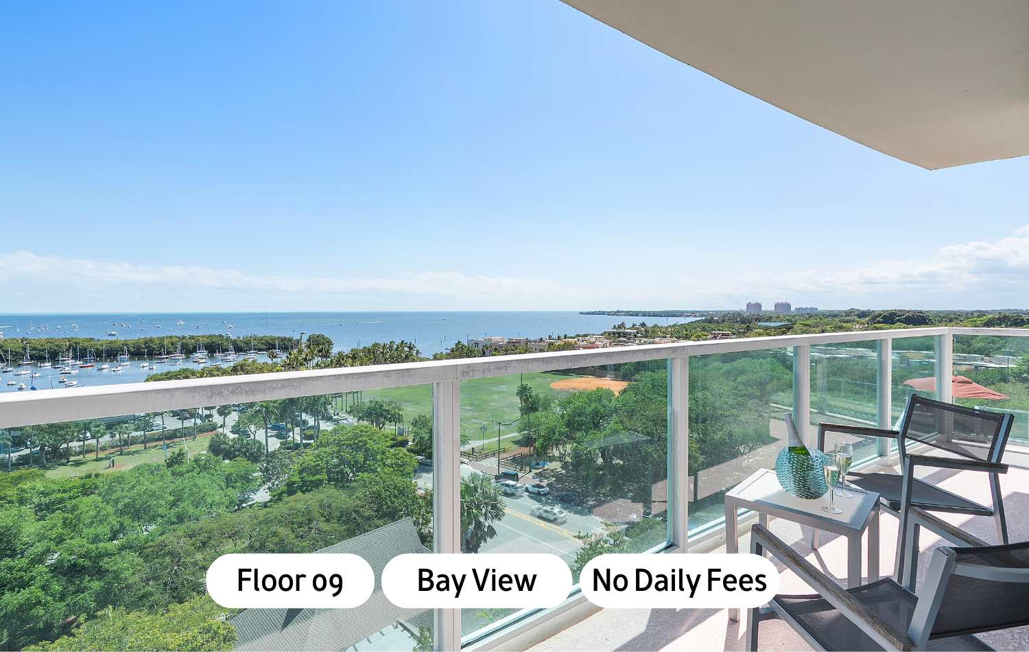 High-floor balcony with a clear glass railing offers panoramic views of a bustling bay filled with boats, a lush green park, and distant cityscapes under a bright blue sky. A small table and two chairs provide comfortable outdoor seating.
