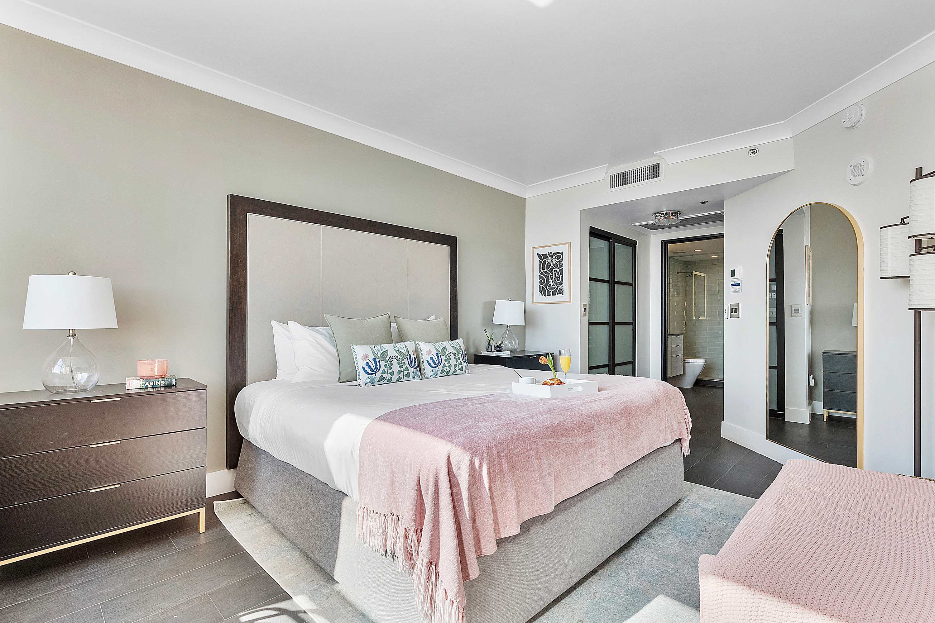 Elegant bedroom featuring a king-sized bed with a tall, upholstered headboard, a dark wood dresser, and a light green accent wall. An ensuite bathroom with a glass shower is visible through a stylish dark-framed doorway.
